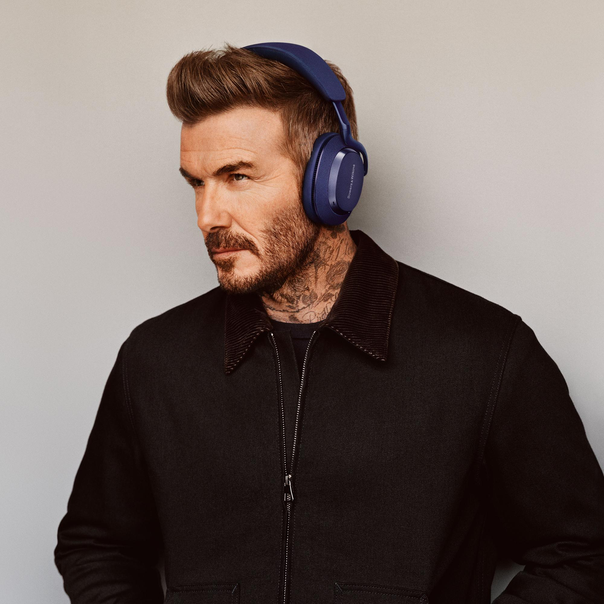 David Beckham trägt blaue Kopfhörer und schaut zur Seite. Er hat Tattoos und eine schwarze Jacke.