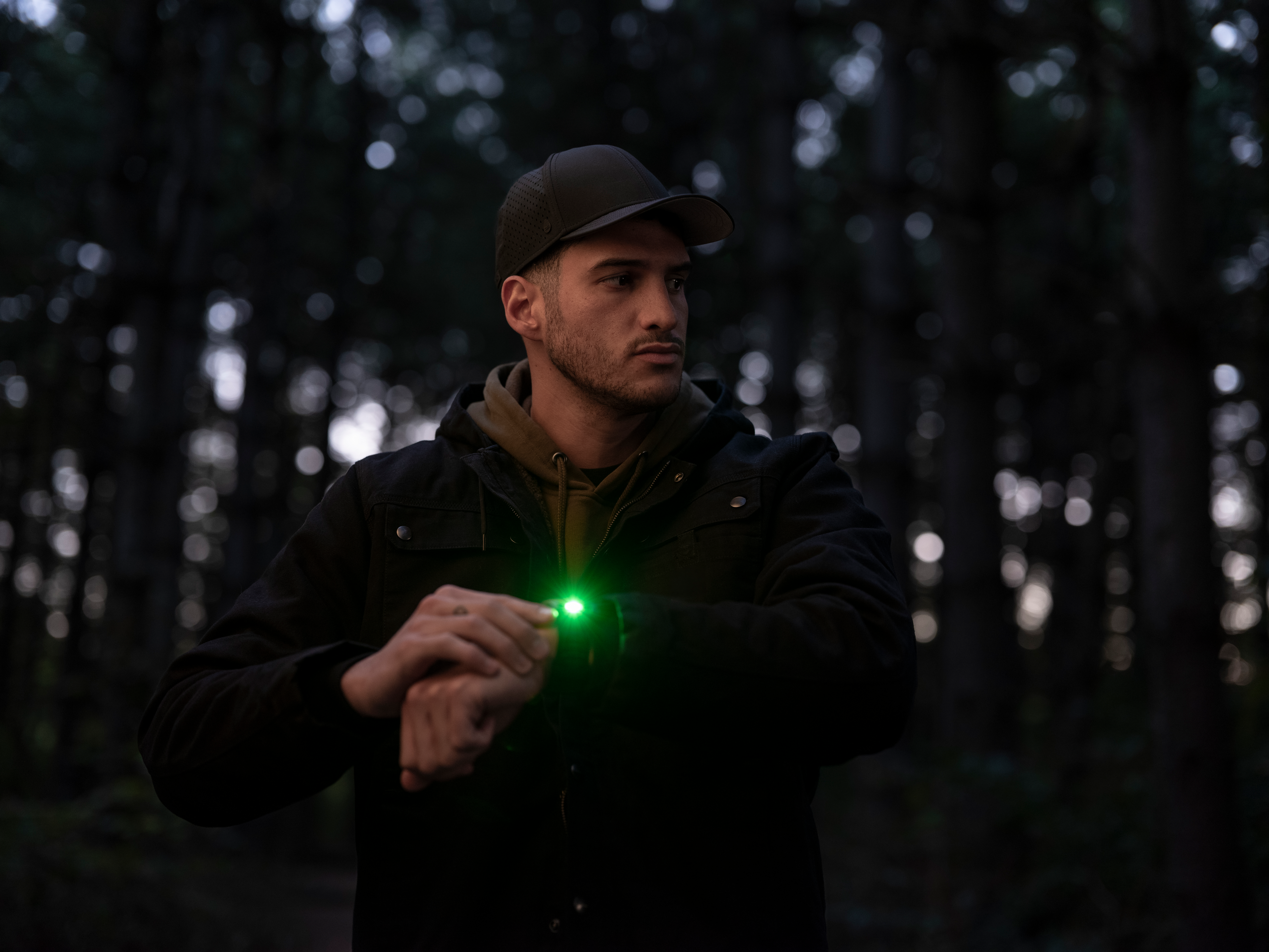 Uomo nel bosco, mano con luce verde, cappellino e giacca scura.