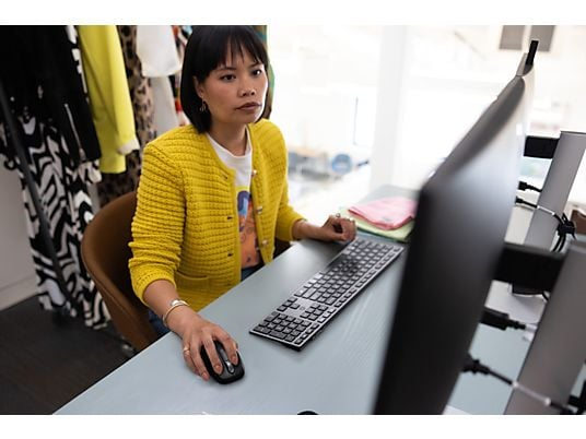 Een vrouw in een gele jas werkt aan een bureau met een computer. Ze gebruikt een muis. Kleren hangen op de achtergrond.
