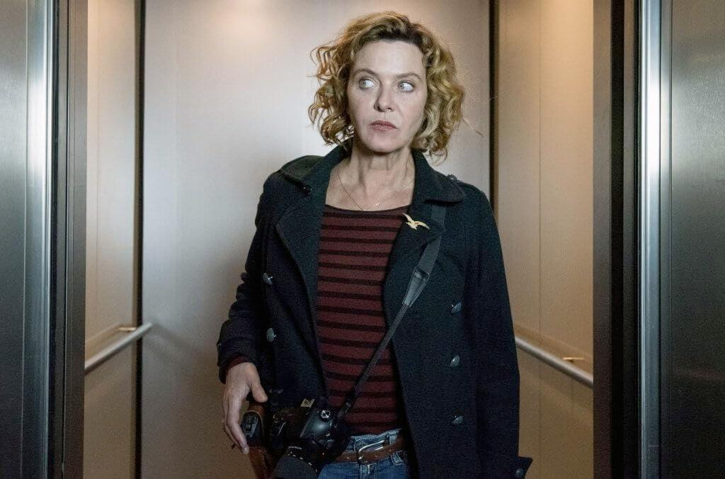 Eine Frau mit lockigem Haar in einem Trenchcoat, die eine Kamera trägt, schaut zur Seite.