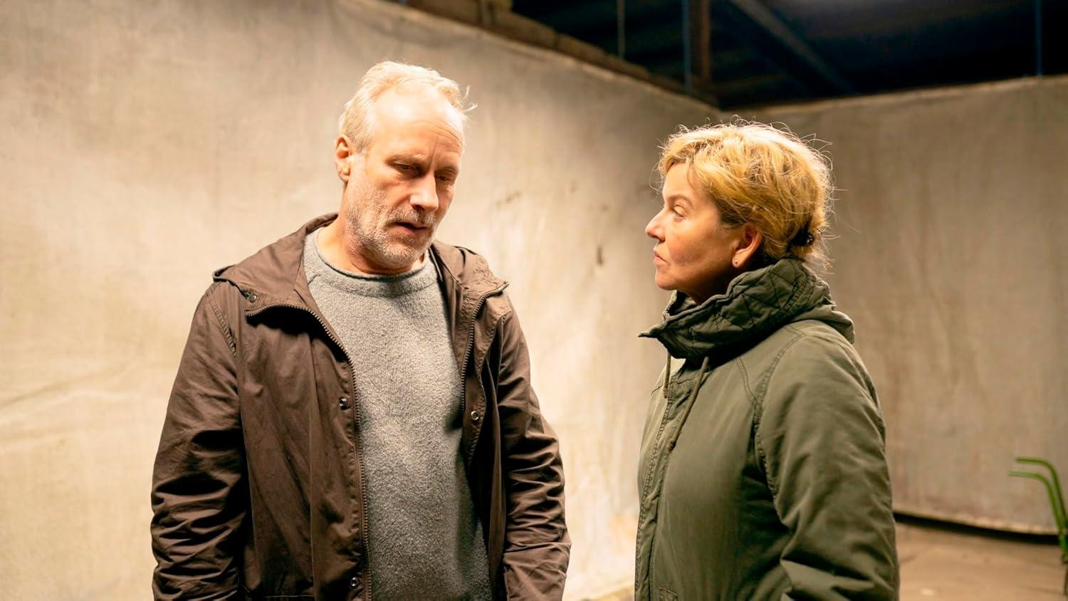 Zwei Personen, ein Mann und eine Frau, stehen in einem Raum mit neutralem Hintergrund und reden.