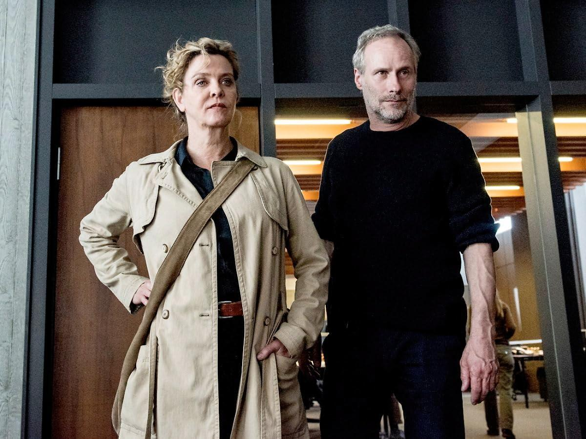Zwei Personen stehen da, schauen nach oben. Eine Frau im Trenchcoat und ein Mann im schwarzen Pullover.