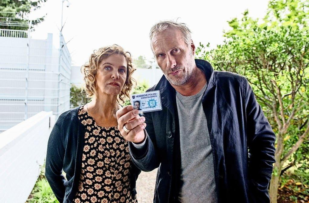 Zwei Personen halten einen Ausweis, in der Nähe eines Zauns und Grün, bei Sonnenschein.