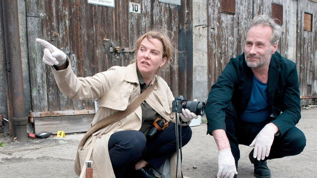 Zwei Detektive am Tatort. Einer zeigt, der andere kniet. Im Hintergrund eine Holzwand.