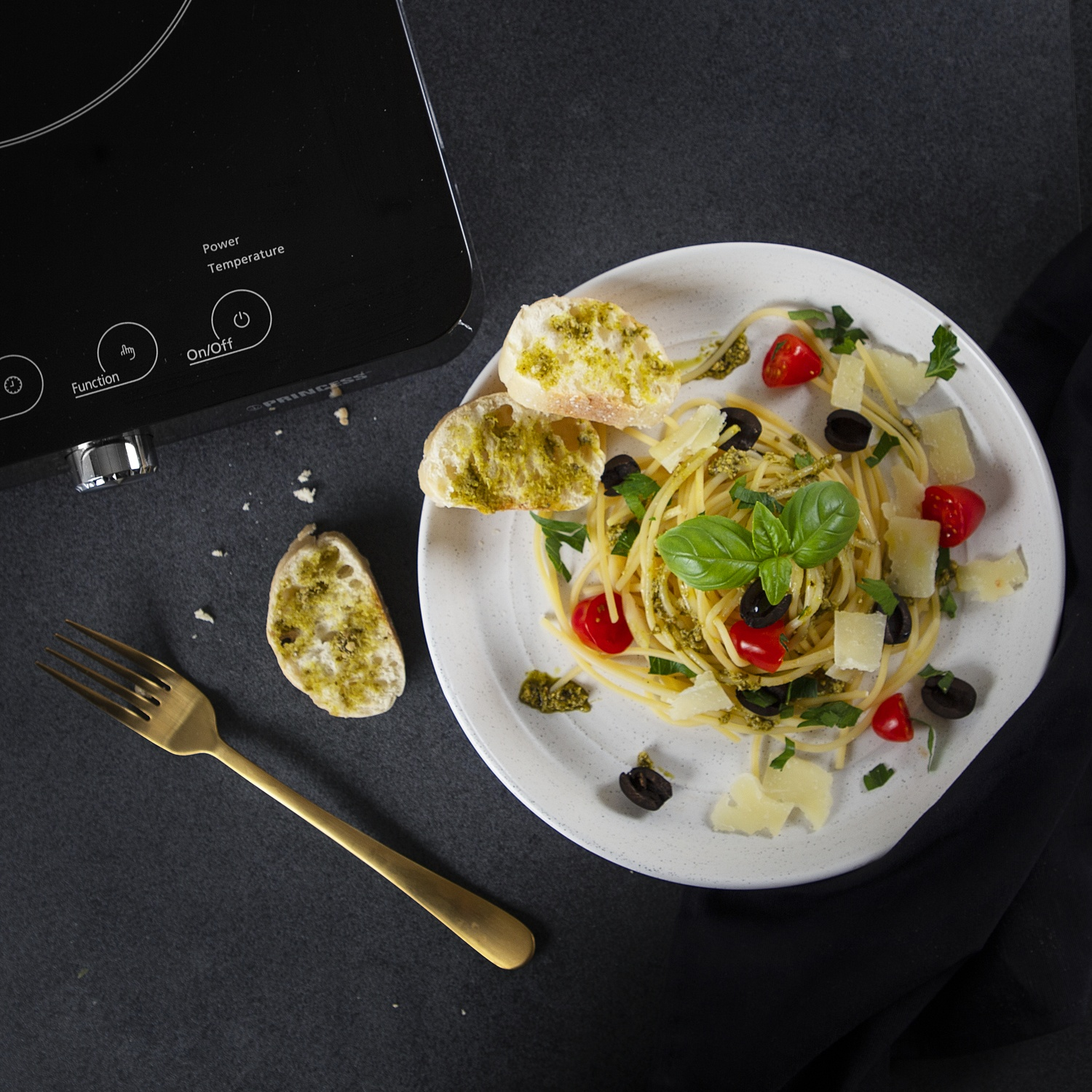 Piatto di pasta con pesto, pomodori e formaggio, con forchetta dorata e fornello nero.