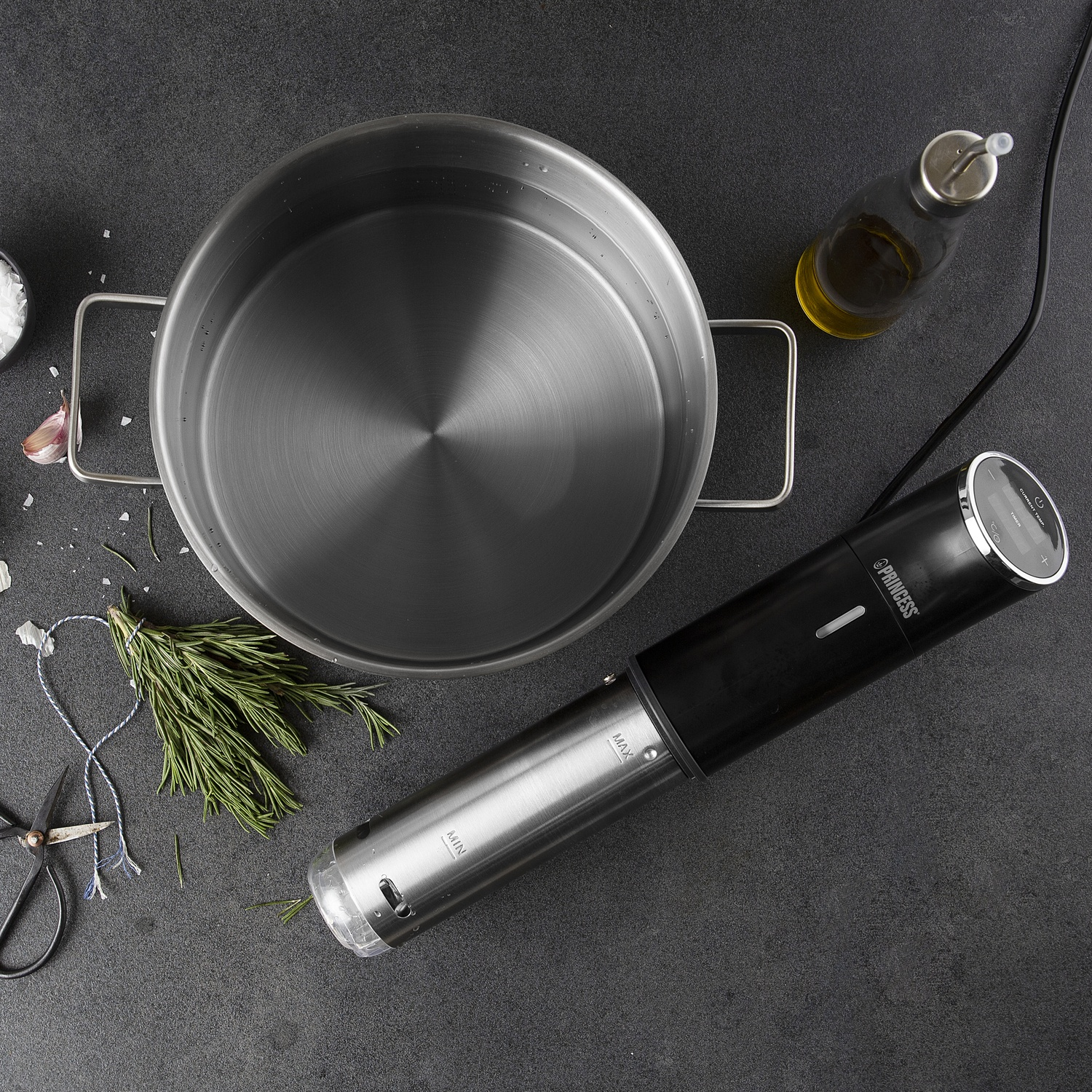 Pentola in acciaio inox con acqua e circolatore a immersione sous vide, accanto erbe e olio oliva.