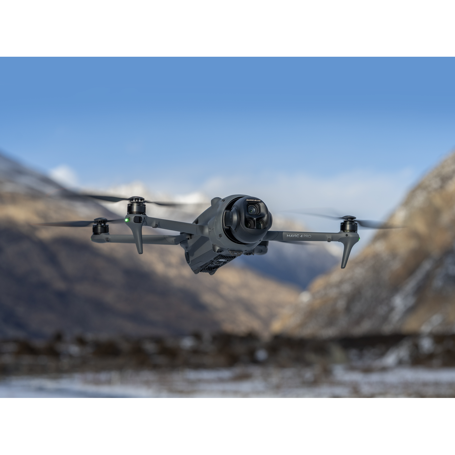A drone flies in front of mountains. The sky is blue and the landscape is covered in snow.