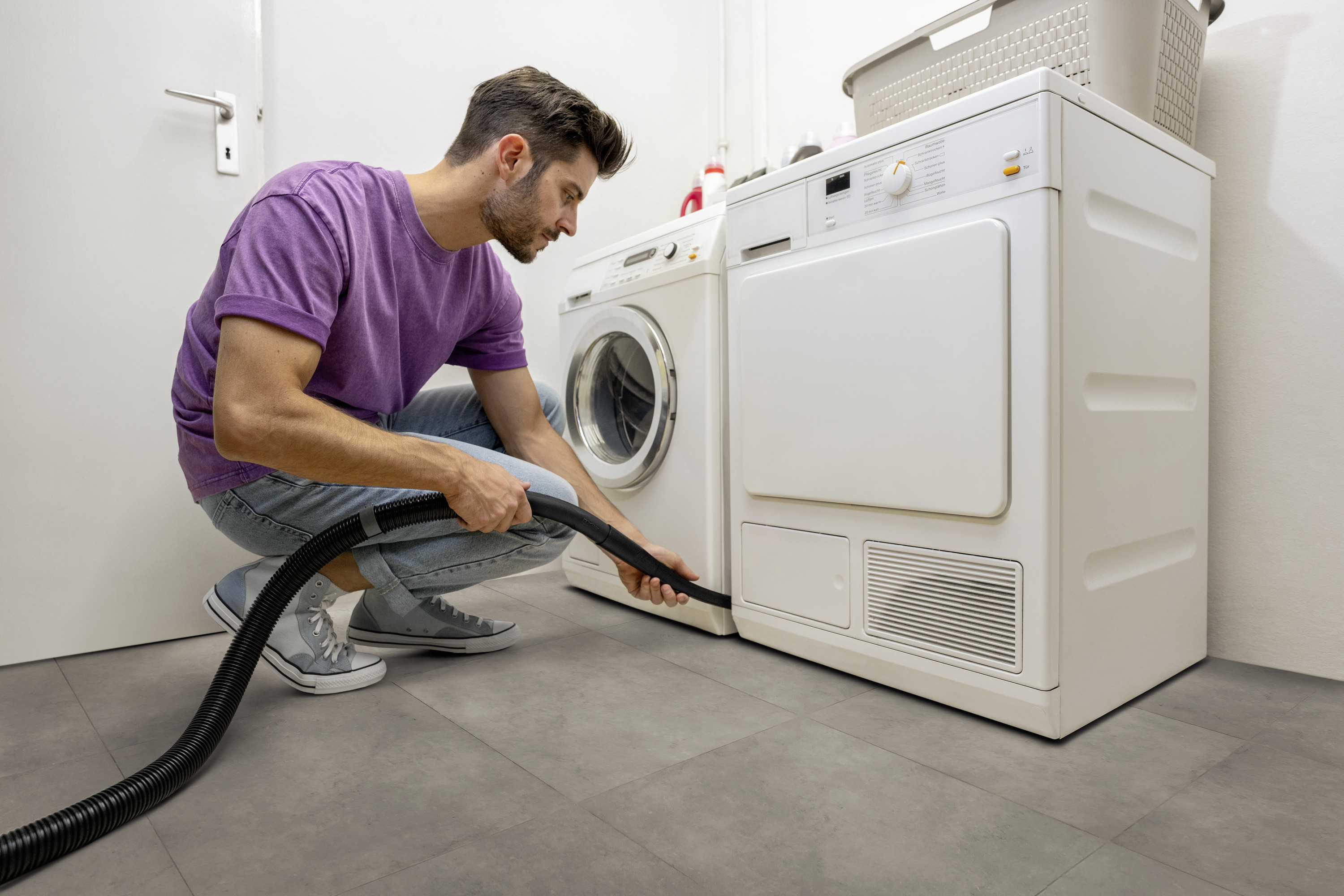 Mężczyzna klęczy i czyści dół pralki za pomocą węża.