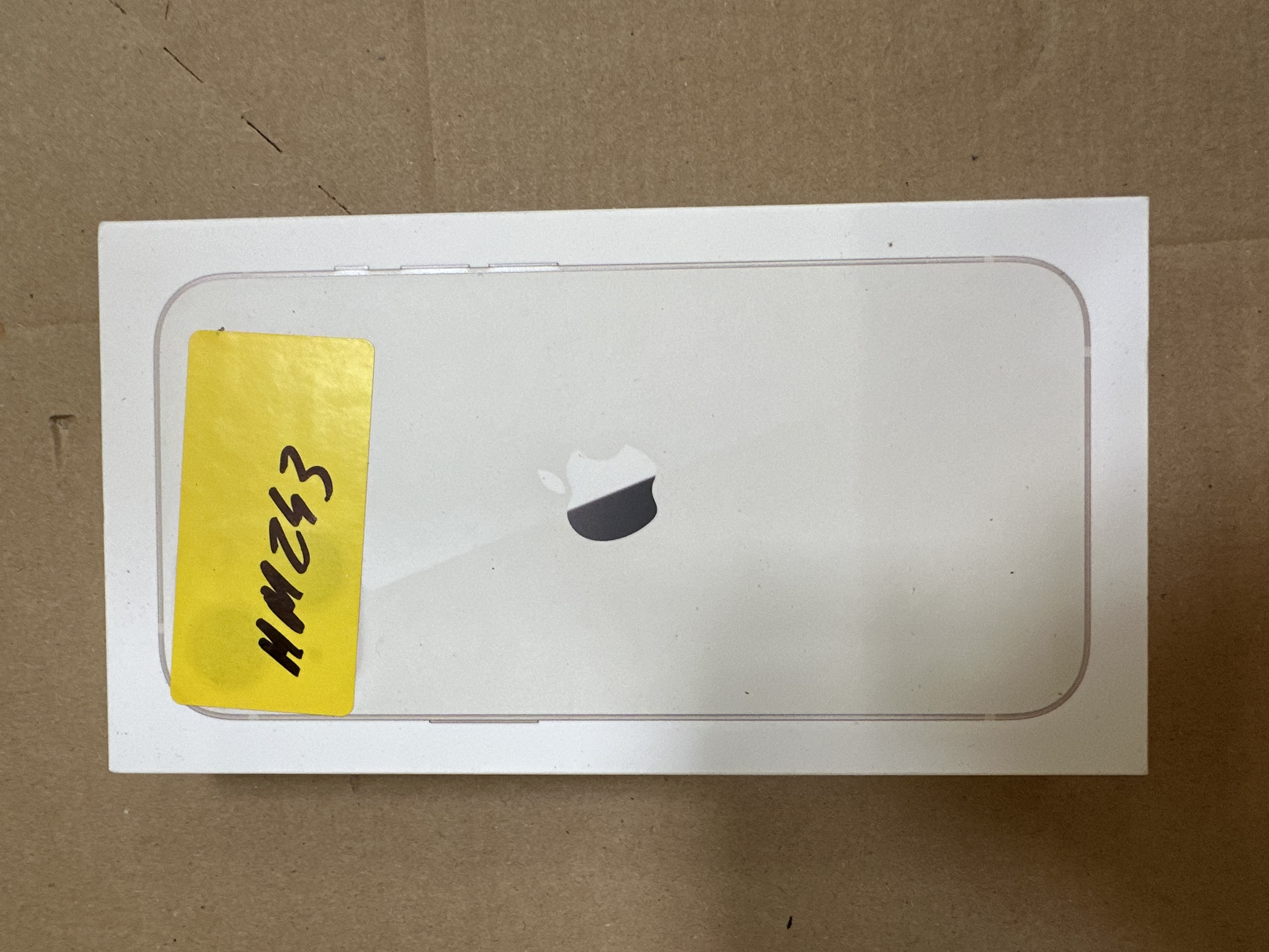Białe pudełko Apple z logo, żółta naklejka, na brązowej powierzchni.