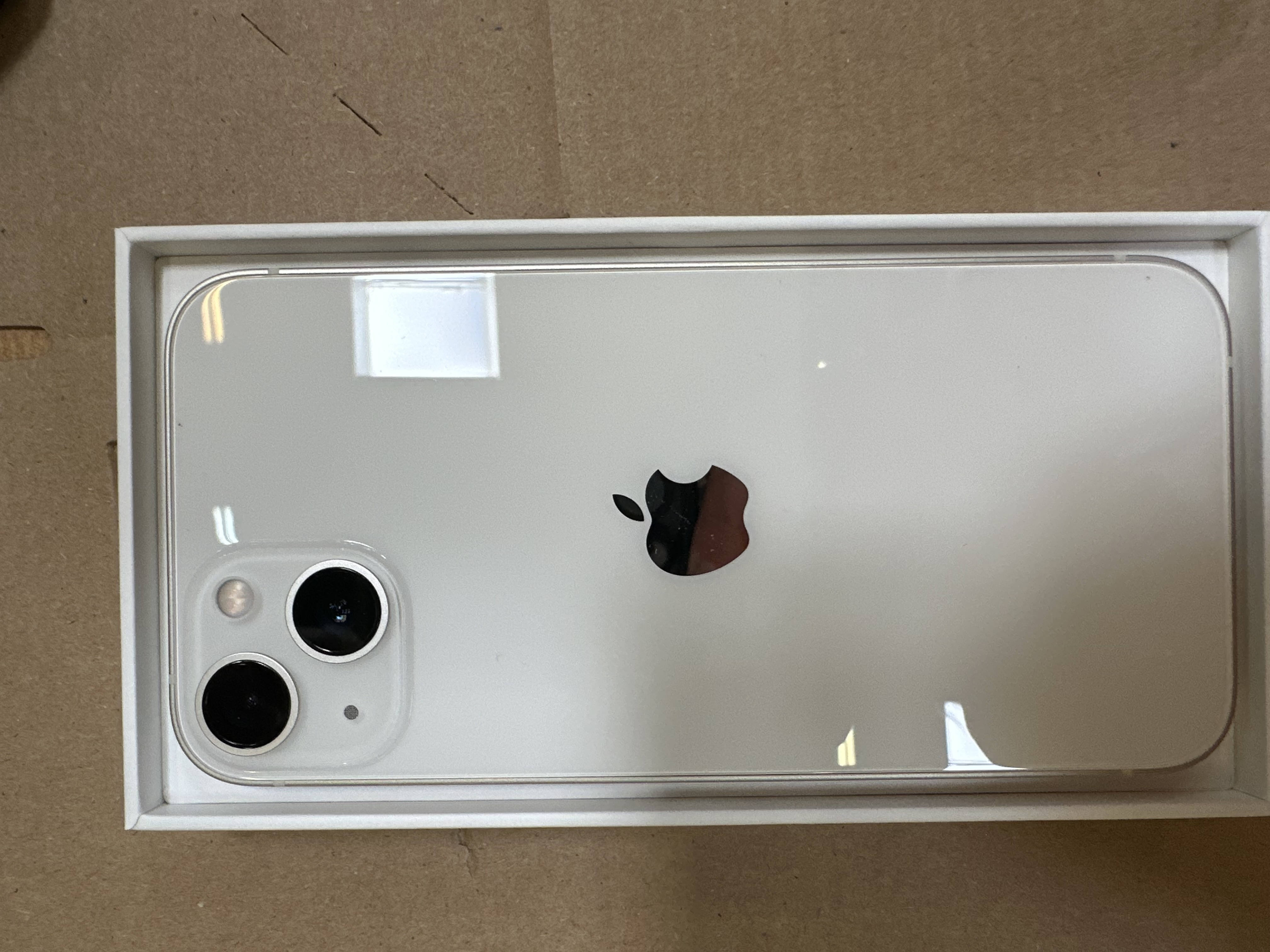 Biały iPhone w białym pudełku, czarne logo Apple, dwa obiektywy, na brązowym kartonie.