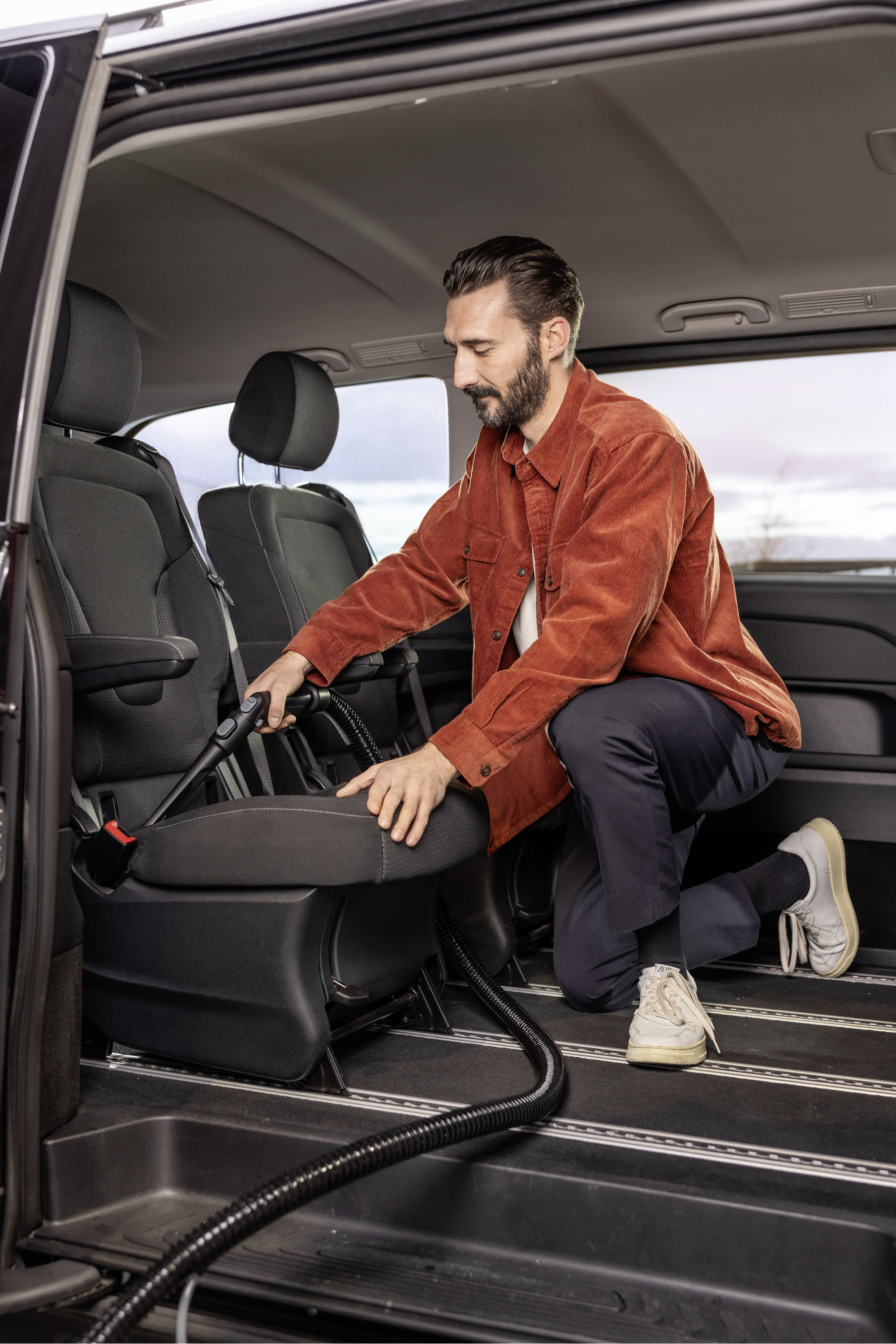 Mężczyzna w pomarańczowej koszuli odkurza wnętrze czarnego samochodu.