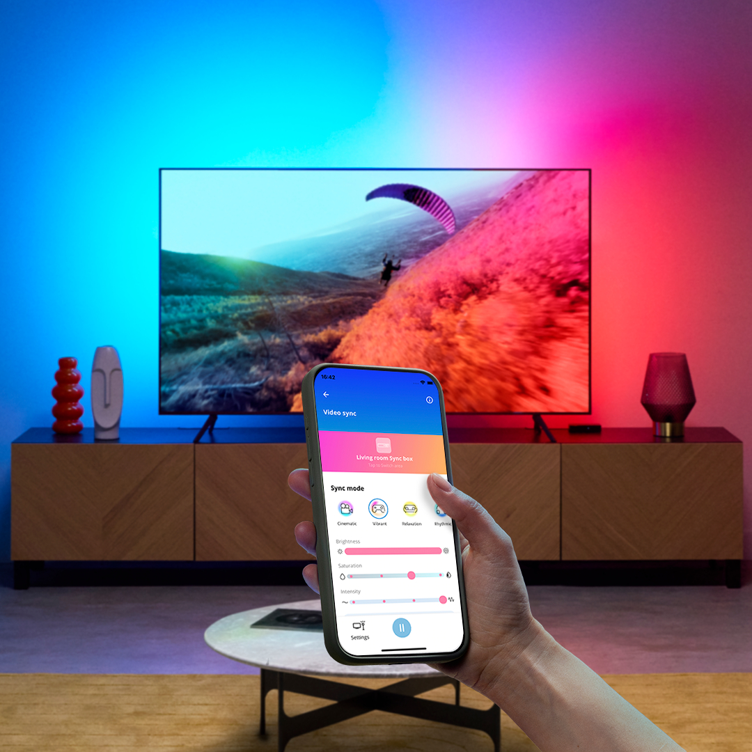 Hand, die ein Telefon mit Smart-Home-App hält, vor TV mit Umgebungsbeleuchtung.