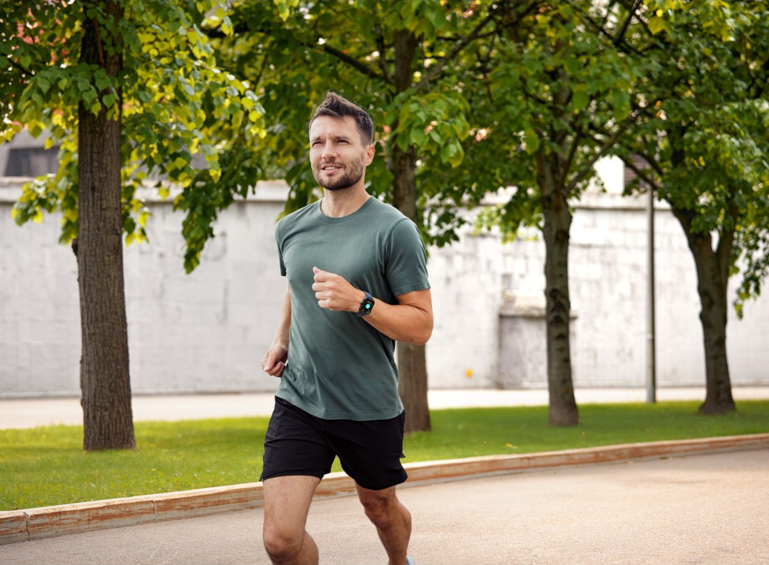 Ein Mann joggt auf einem Weg in der Nähe von Bäumen. Er trägt ein grünes Hemd und schwarze Shorts. An seinem Handgelenk ist eine Smartwatch.