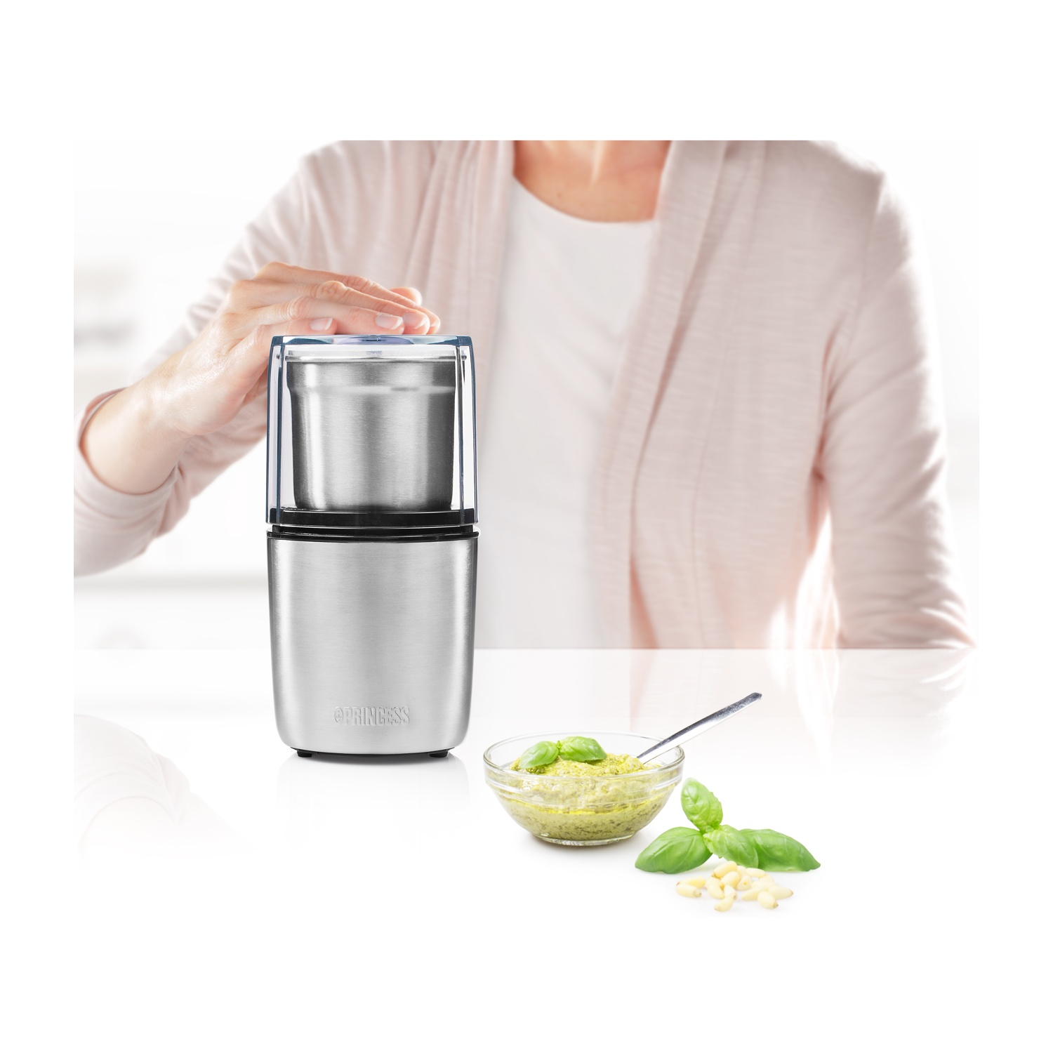 Una donna usa un macinacaffè elettrico argentato. Una ciotola di pesto e ingredienti sul tavolo.