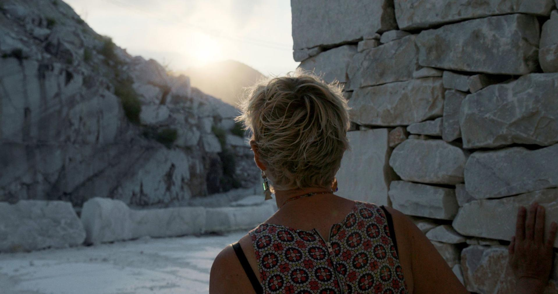 Frau mit blonden Haaren, die eine Steinmauer betrachtet, helles Sonnenlicht.