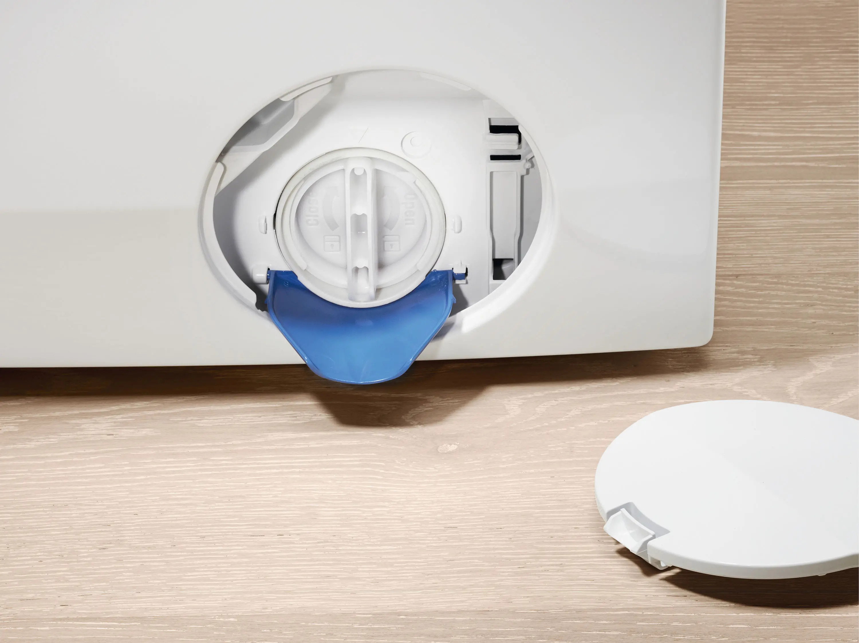 Zbliżenie pralki z otwartą pokrywą filtra, pokazującą niebieski uchwyt i białą pokrywę na drewnianej podłodze.