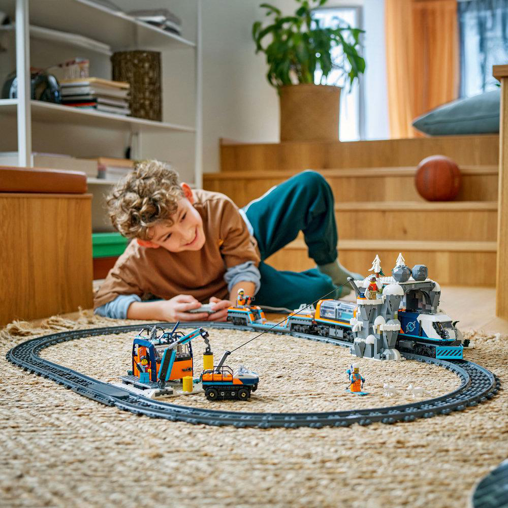 Ein Junge spielt mit einem Spielzeugeisenbahnset auf einem Teppich, umgeben von Möbeln und Treppen.