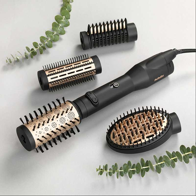 Uno styler per capelli nero con vari accessori e foglie verdi su uno sfondo grigio.