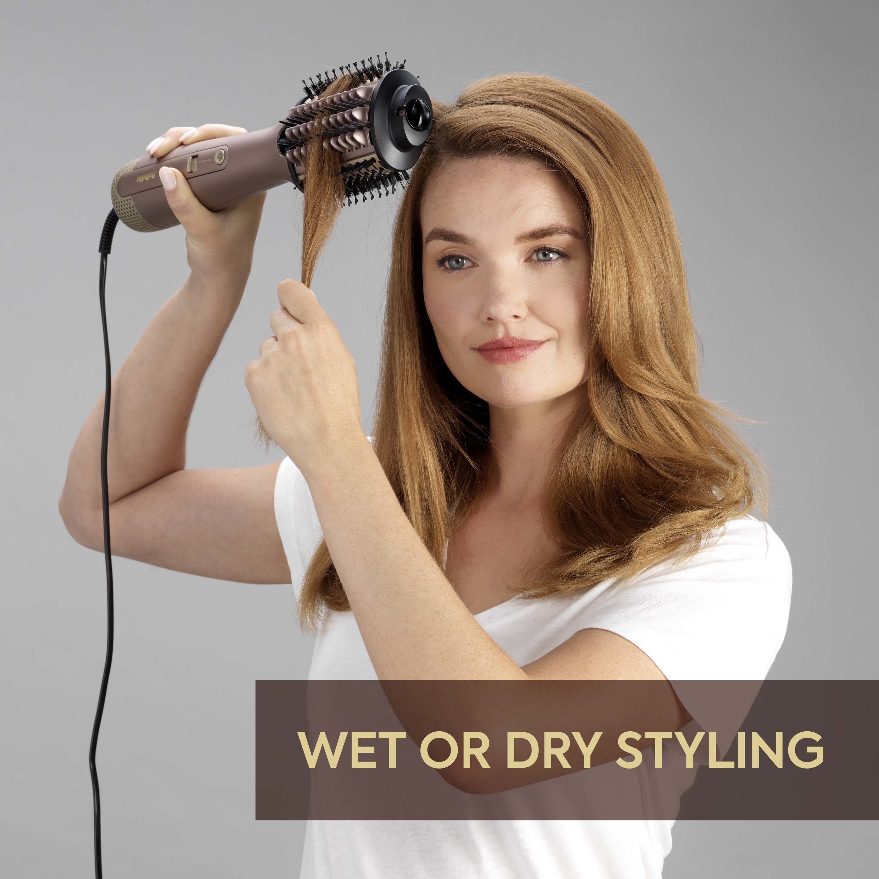 Donna con capelli ramati si acconcia con una spazzola ad aria calda color oro rosa su sfondo grigio.