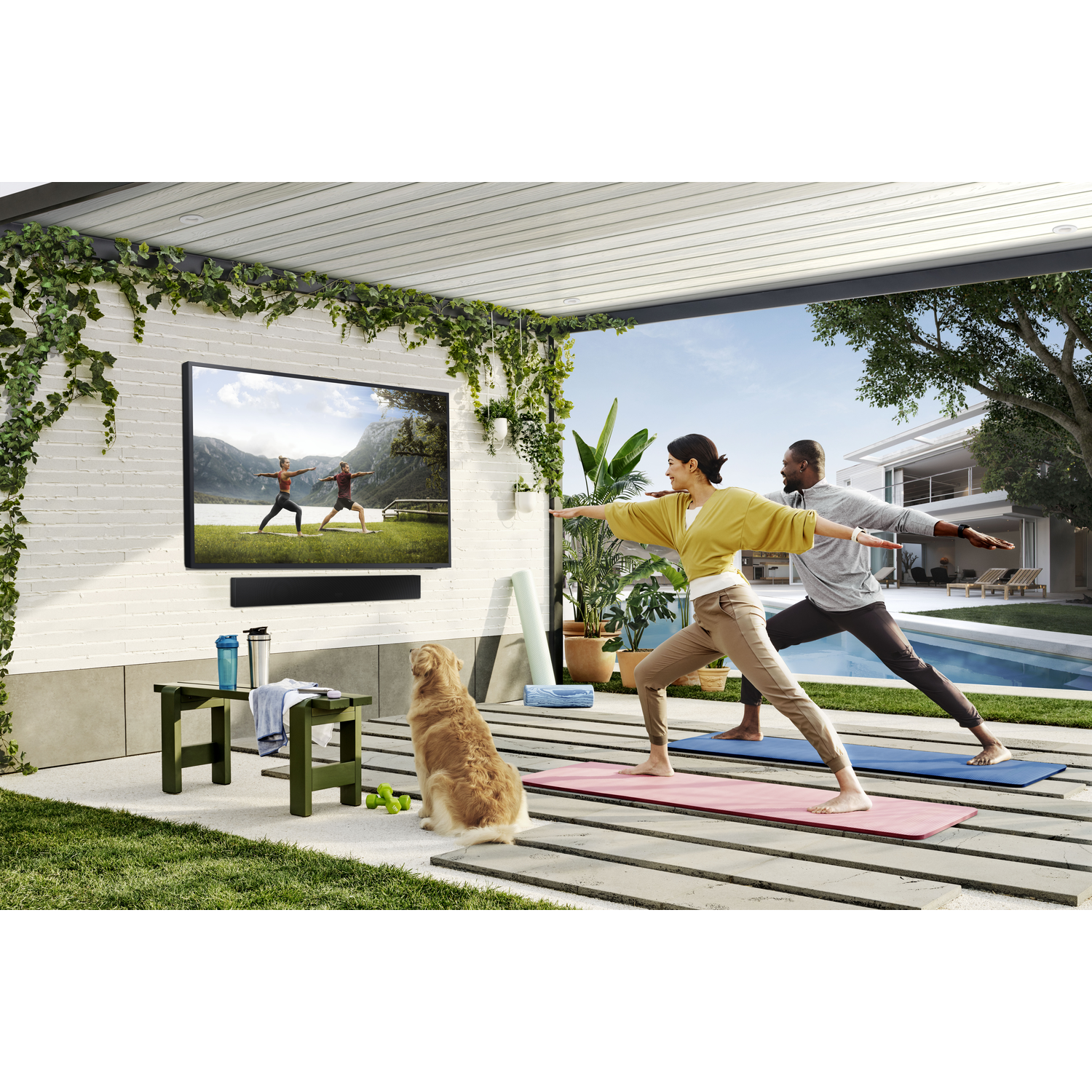 A couple does yoga outdoors on mats, with a TV and dog nearby.