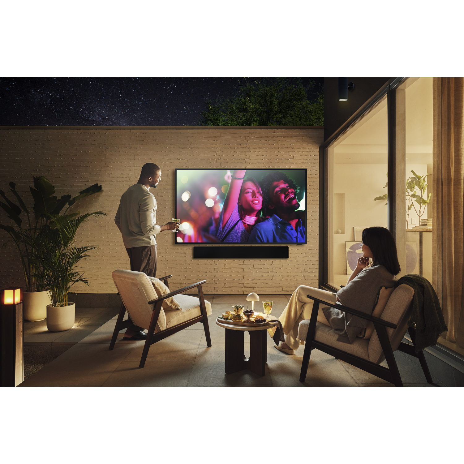 A couple relaxes outdoors, watching a vibrant concert on a TV.