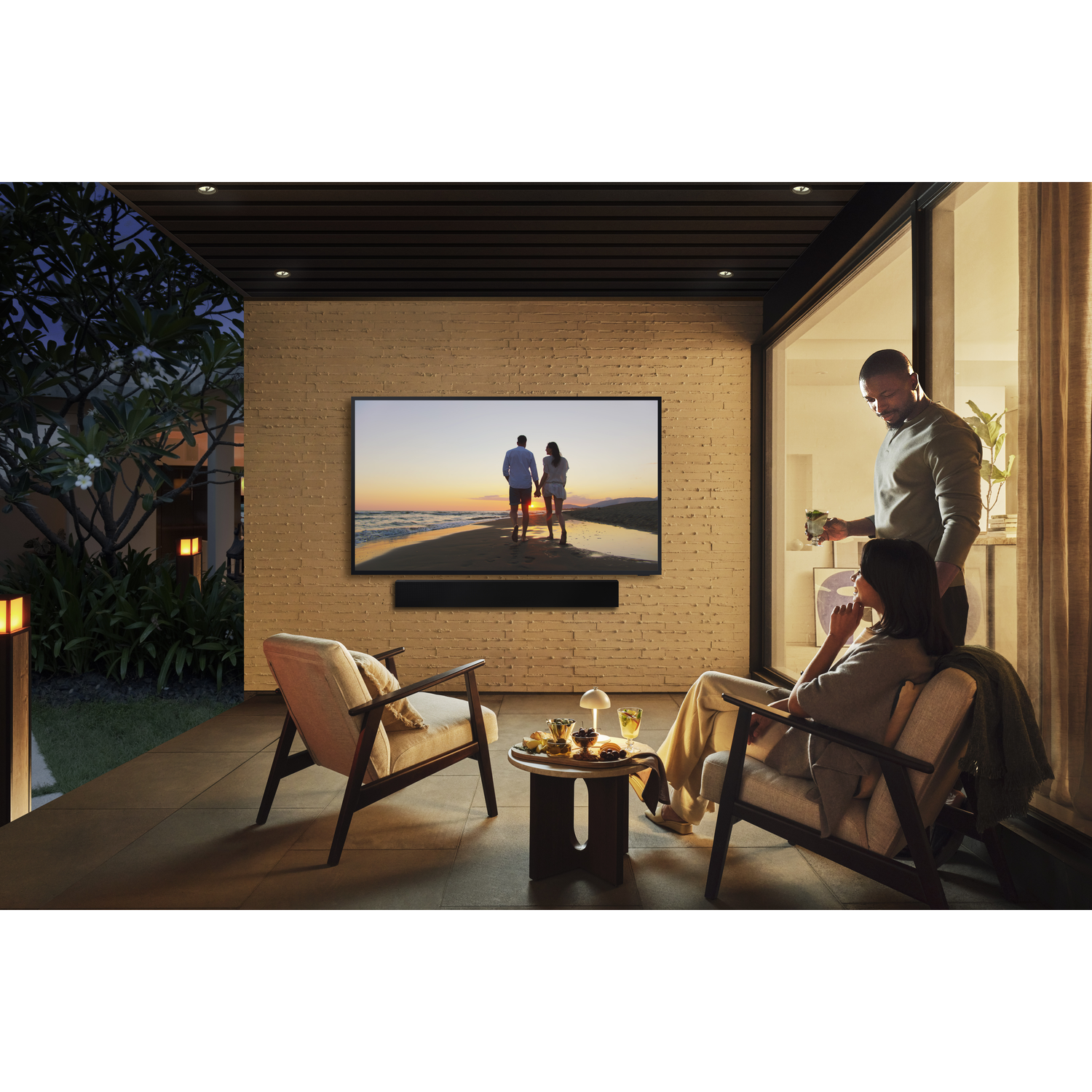 Couple relaxing on patio, TV shows beach scene, man serves drinks. Cozy atmosphere with outdoor lighting.