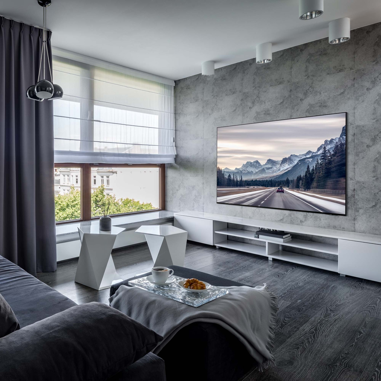 Soggiorno con divano grigio, tavoli bianchi e TV che mostra una strada di montagna.
