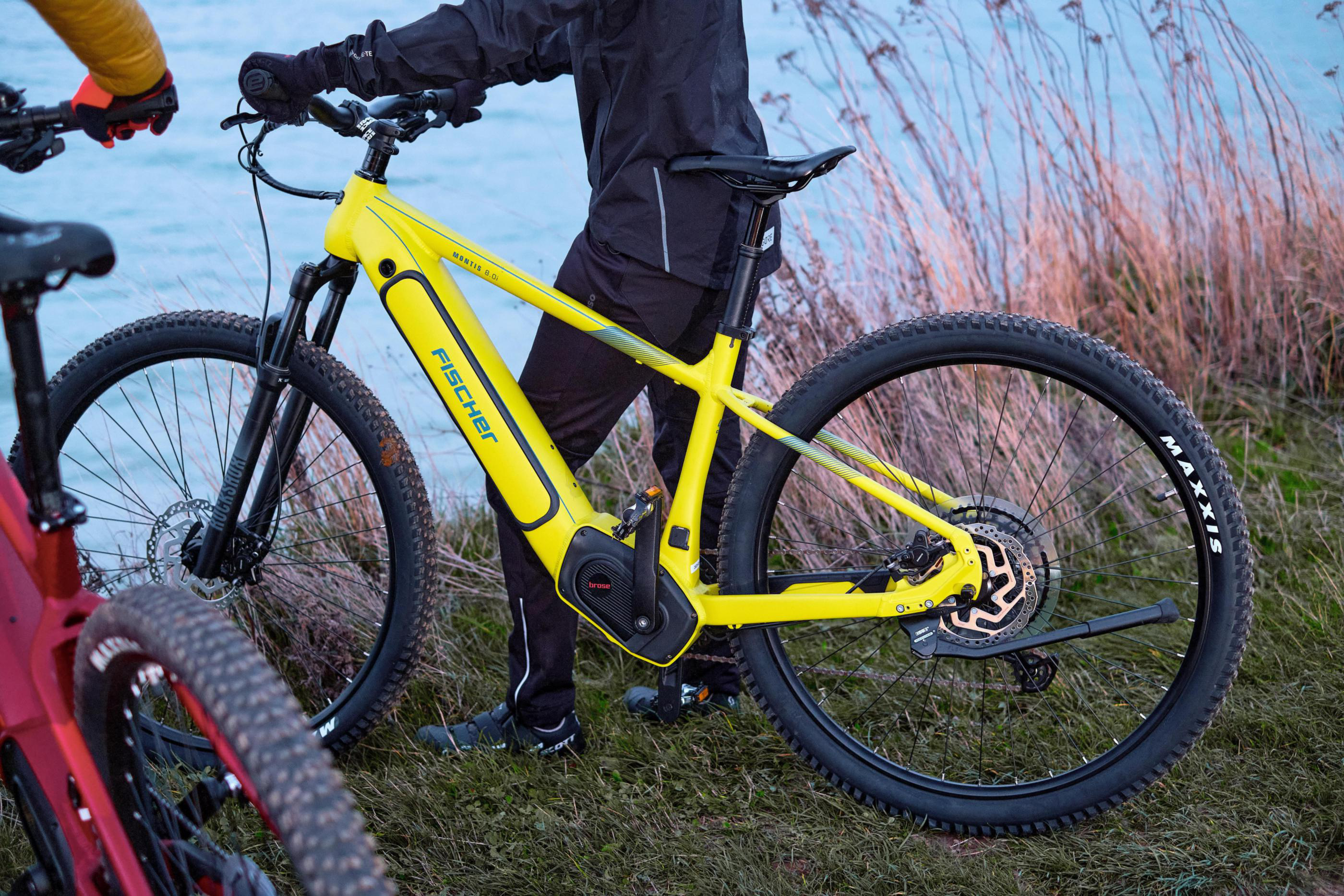 Gelbes Fischer-E-Bike steht auf Gras mit einer Person in Schwarz. Im Hintergrund befindet sich ein Gewässer.