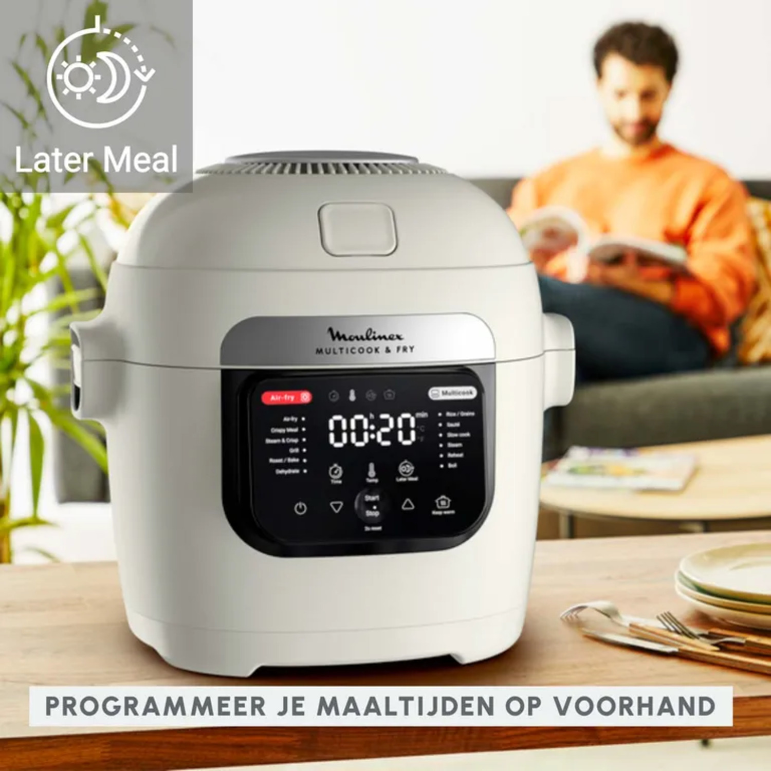 White Moulinex multicooker with digital display on a wooden table, man reading in background.