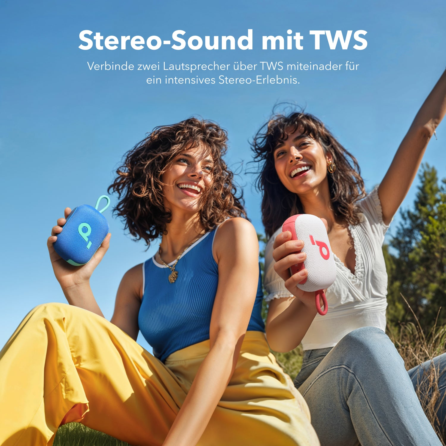 Zwei lachende Frauen sitzen draußen und halten blaue und weiße Lautsprecher in der Hand.