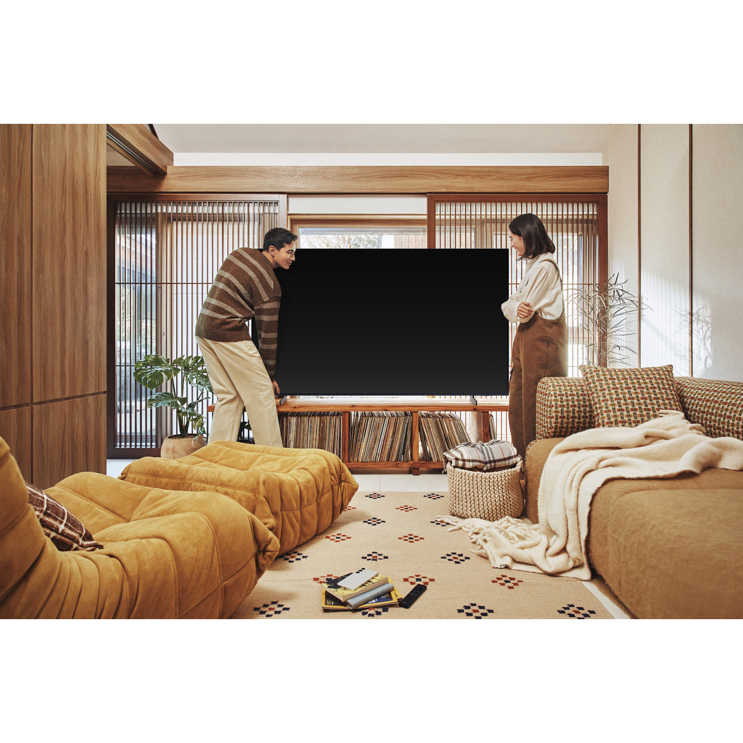 A couple places a large black TV in a living room featuring brown furniture and wood accents.