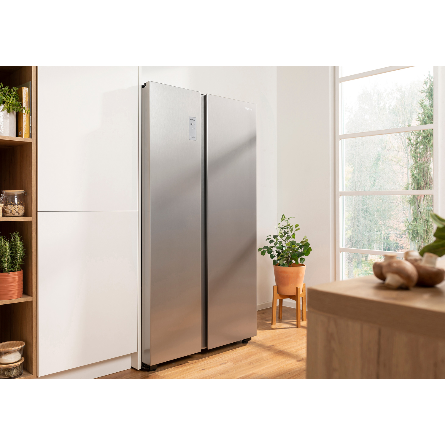 A stainless steel refrigerator stands in a bright kitchen next to white cabinets and plants.