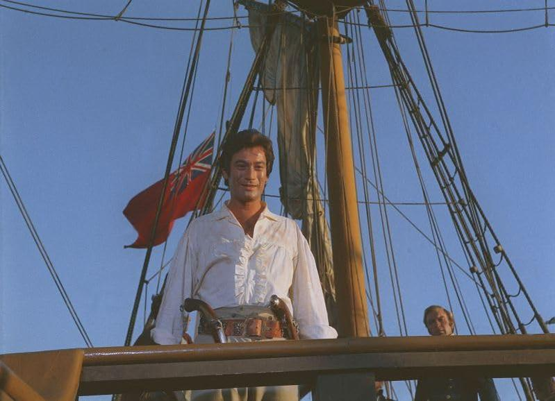 Ein Mann auf einem Schiff, mit Pistolen am Gürtel, vor einer Flagge mit rotem Hintergrund.