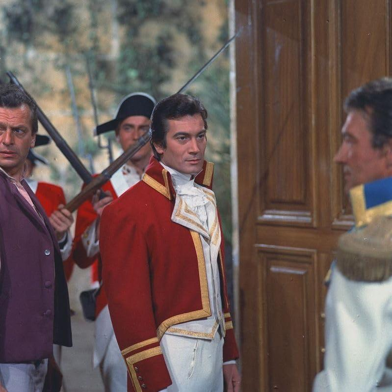 Szene mit Männern in historischen Uniformen, einer mit roter Jacke und Goldbesatz, im Freien.
