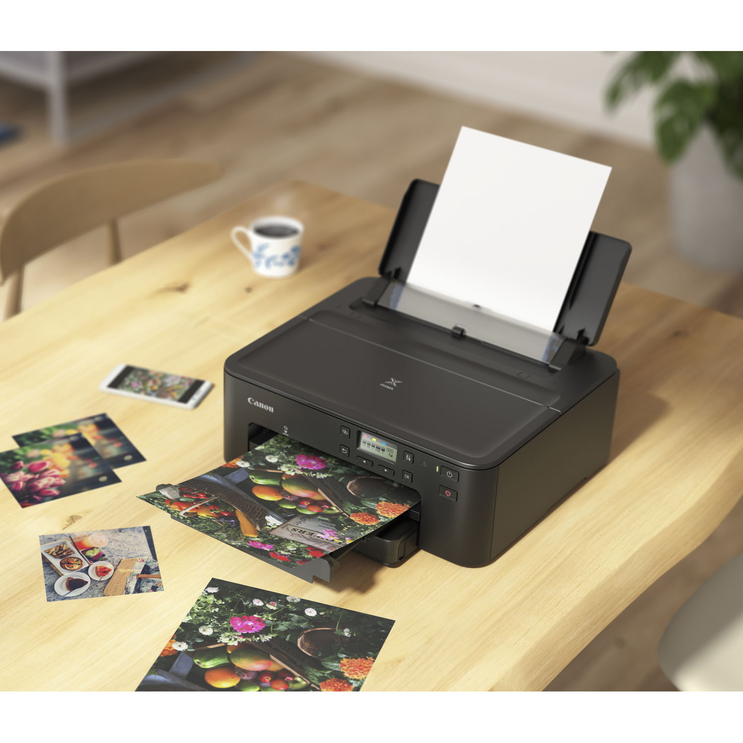 A black Canon printer prints photos onto a wooden table. Printed photos and a phone are scattered. A cup of coffee sits nearby.