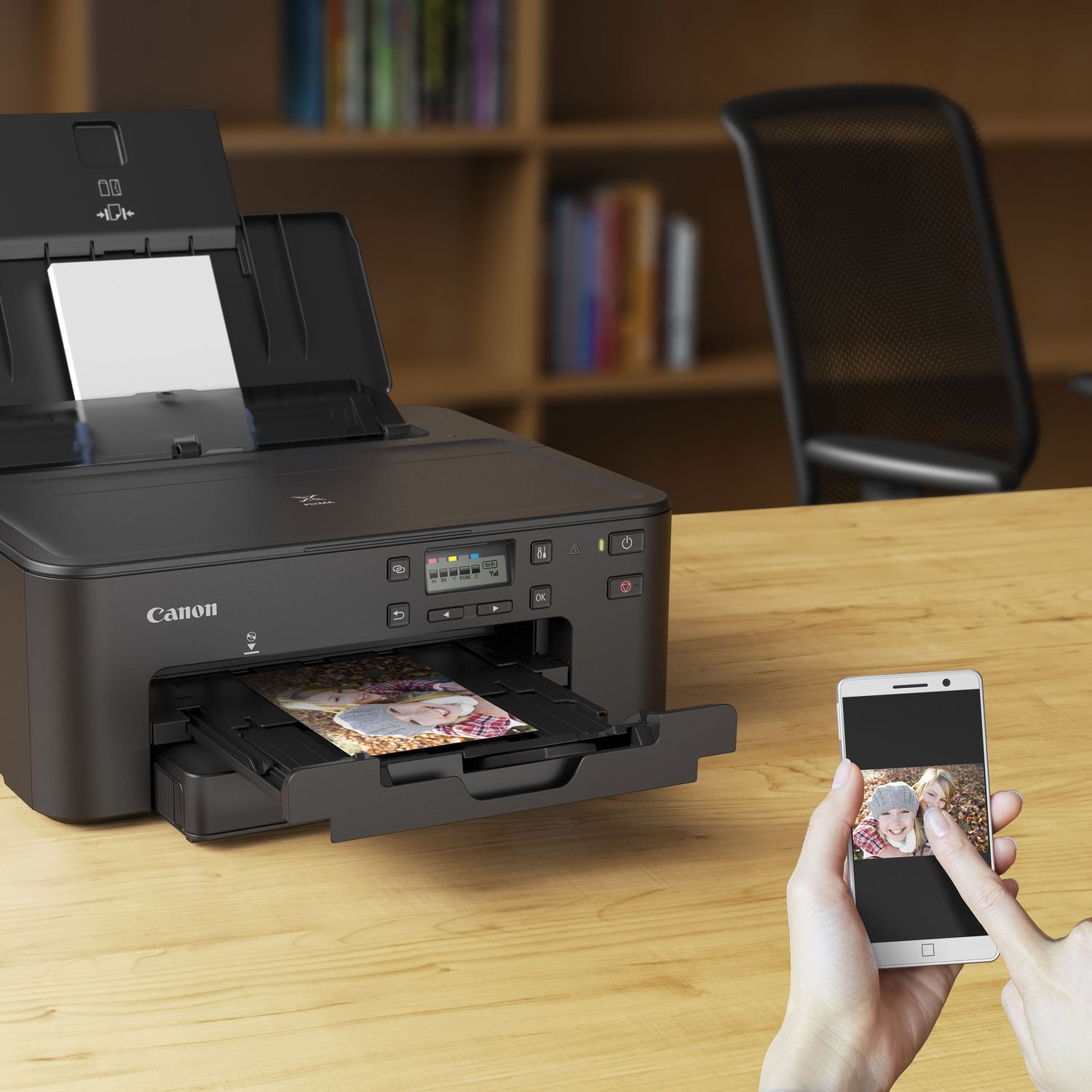 A black Canon printer with a photo in the tray. A smartphone displays the same photo. A hand is touching the phone. Bookshelf in background.