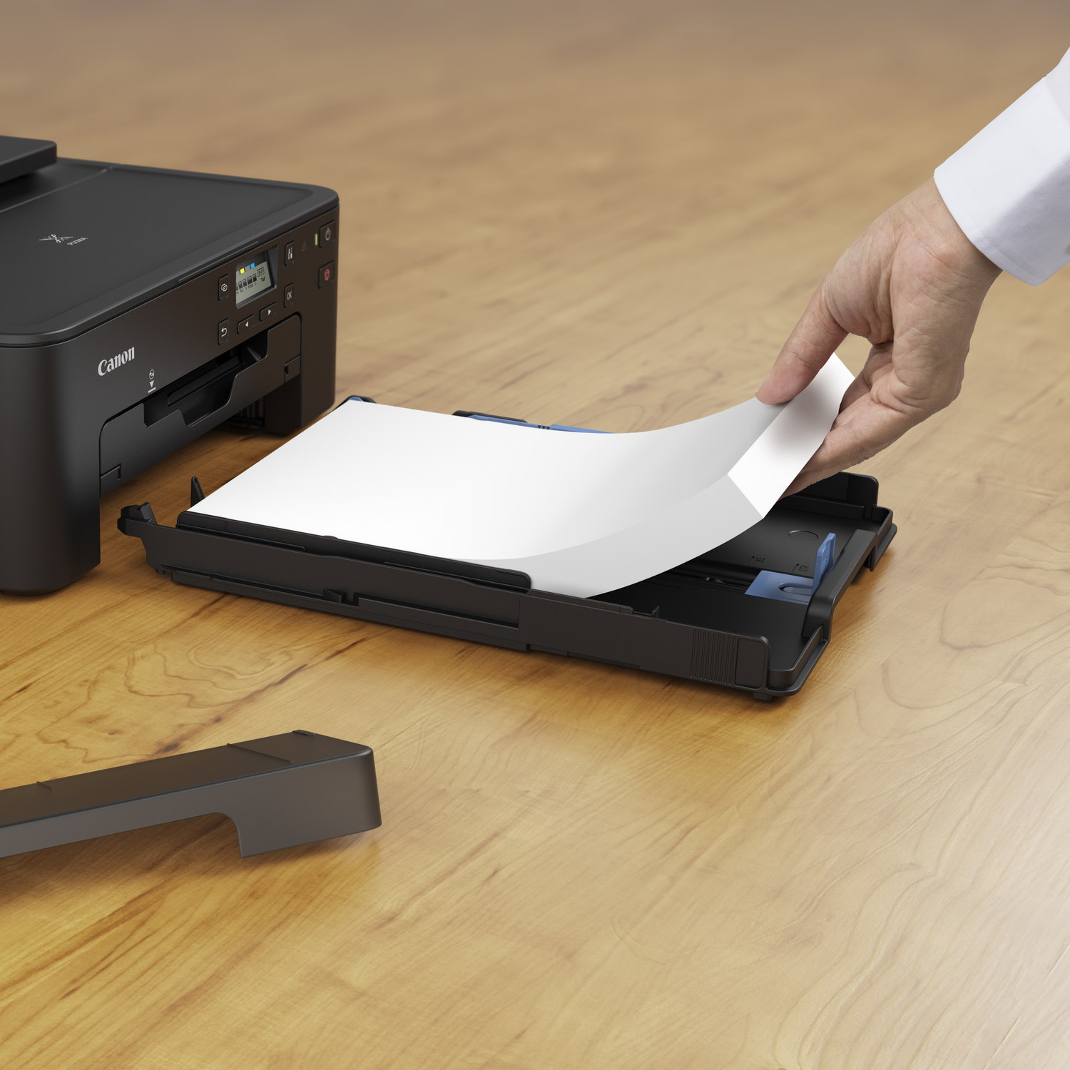 A hand inserts paper into a black Canon printer's paper tray. The printer sits on a wooden surface, with a detached tray cover nearby.