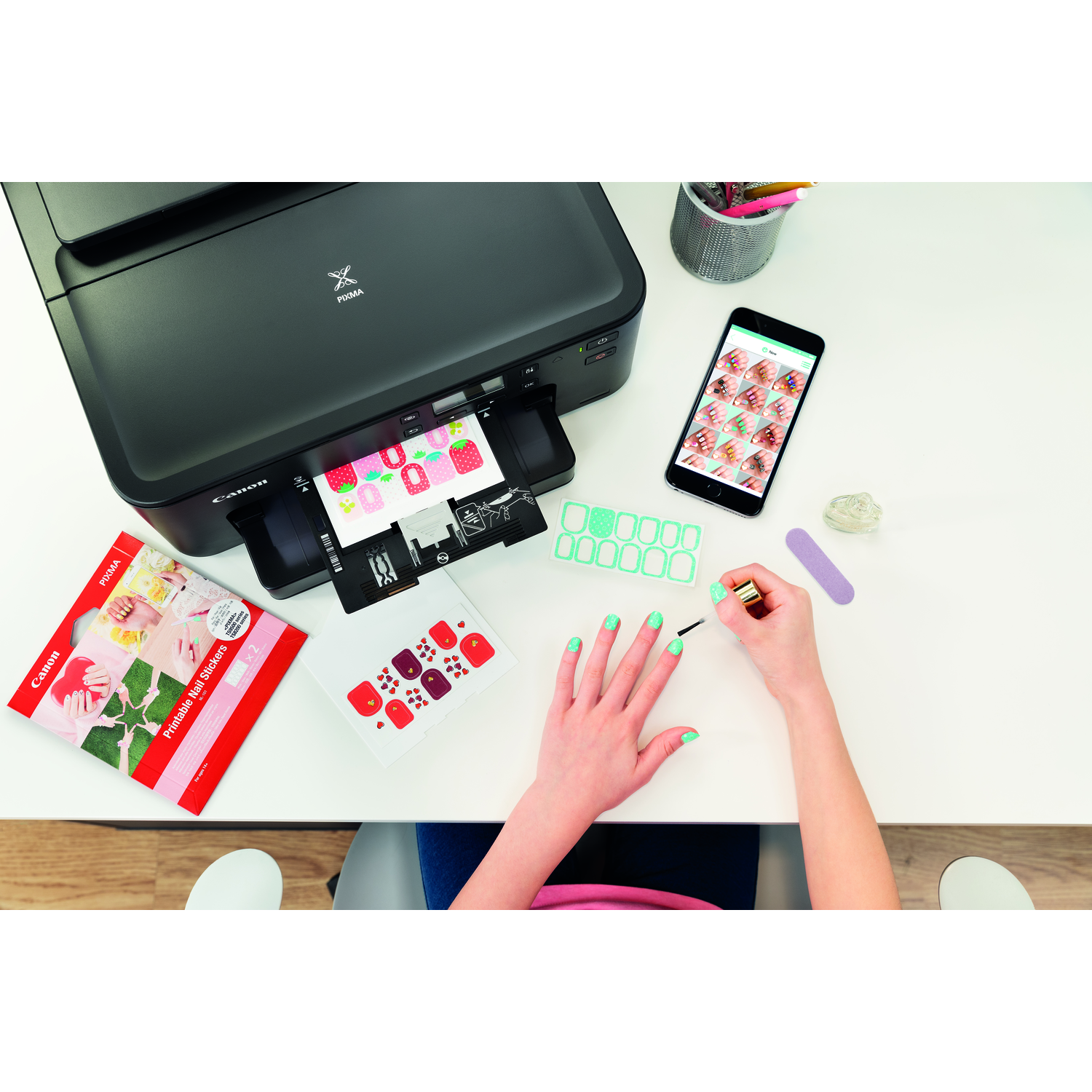 Printer printing stickers. A person is painting their nails. A smartphone with sticker designs is visible.