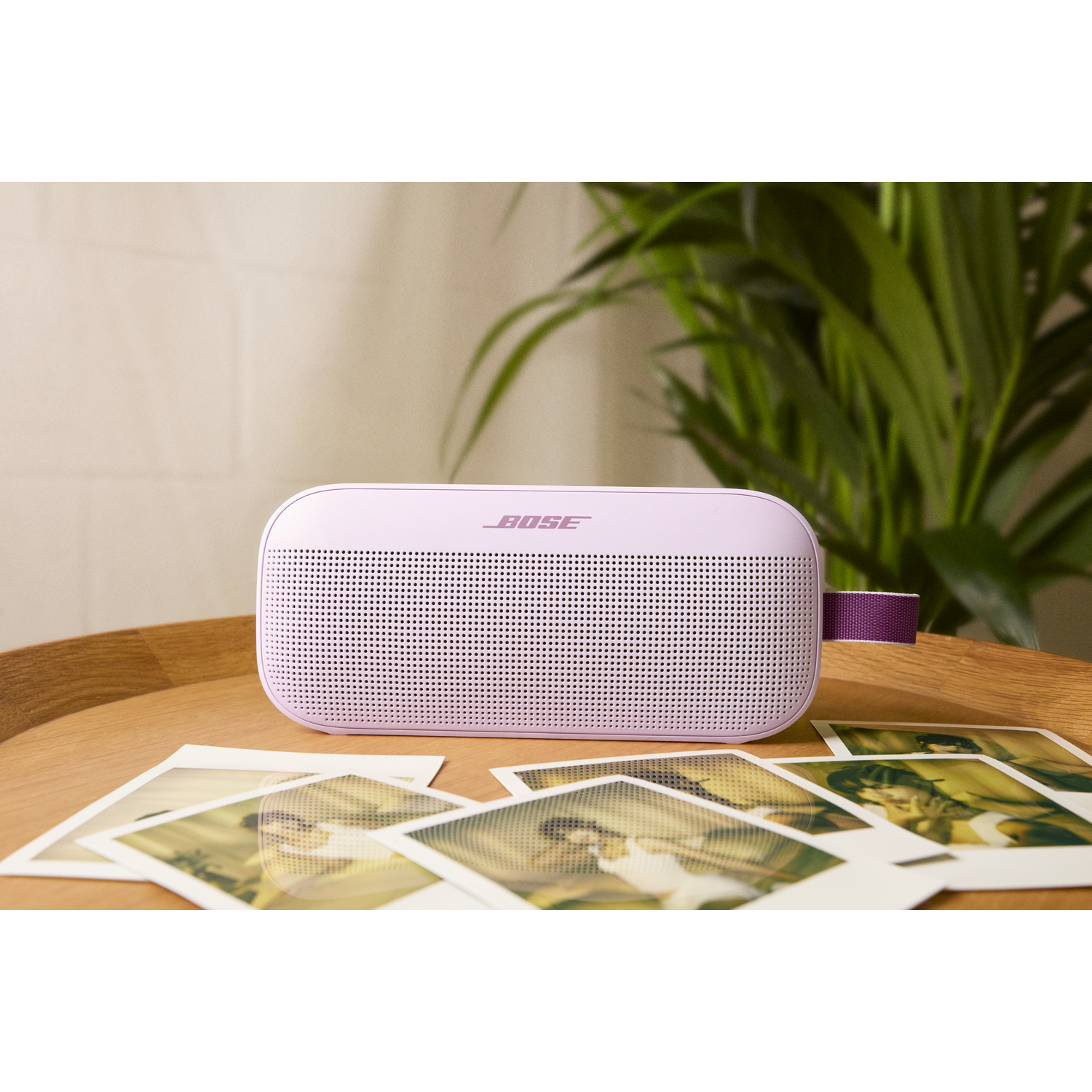 Een roze Bose-luidspreker staat op een houten tafel met polaroidfoto's. Groene plant op de achtergrond. De luidspreker heeft een paars handvat.