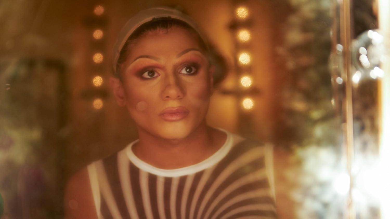 Nahaufnahme einer Person mit Make-up, trägt ein gestreiftes Oberteil, blickt leicht aus der Kamera.