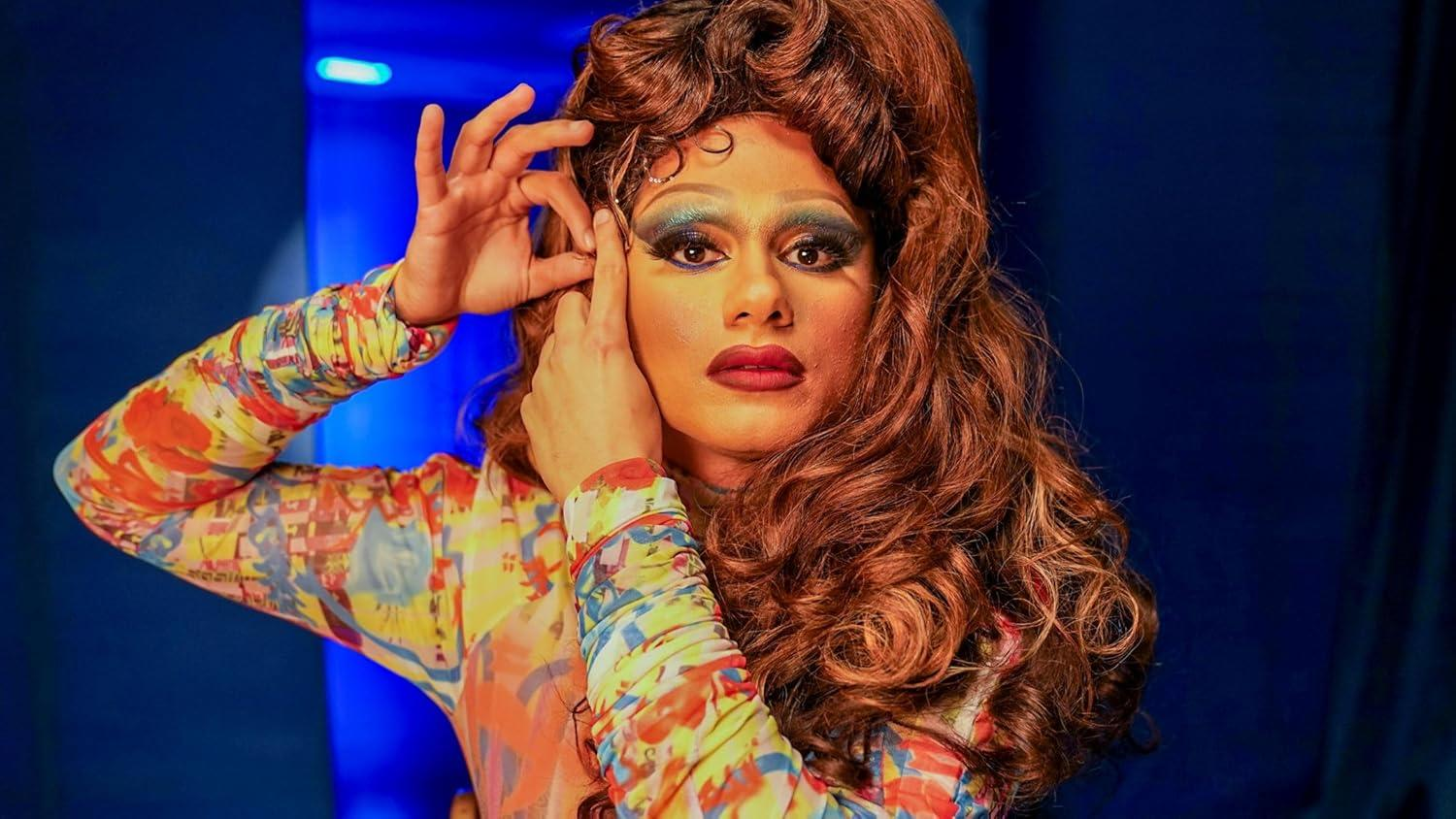 Eine Person mit Make-up und Perücke posiert, Hände nahe am Gesicht. Sie trägt ein buntes Oberteil vor blauem Hintergrund.