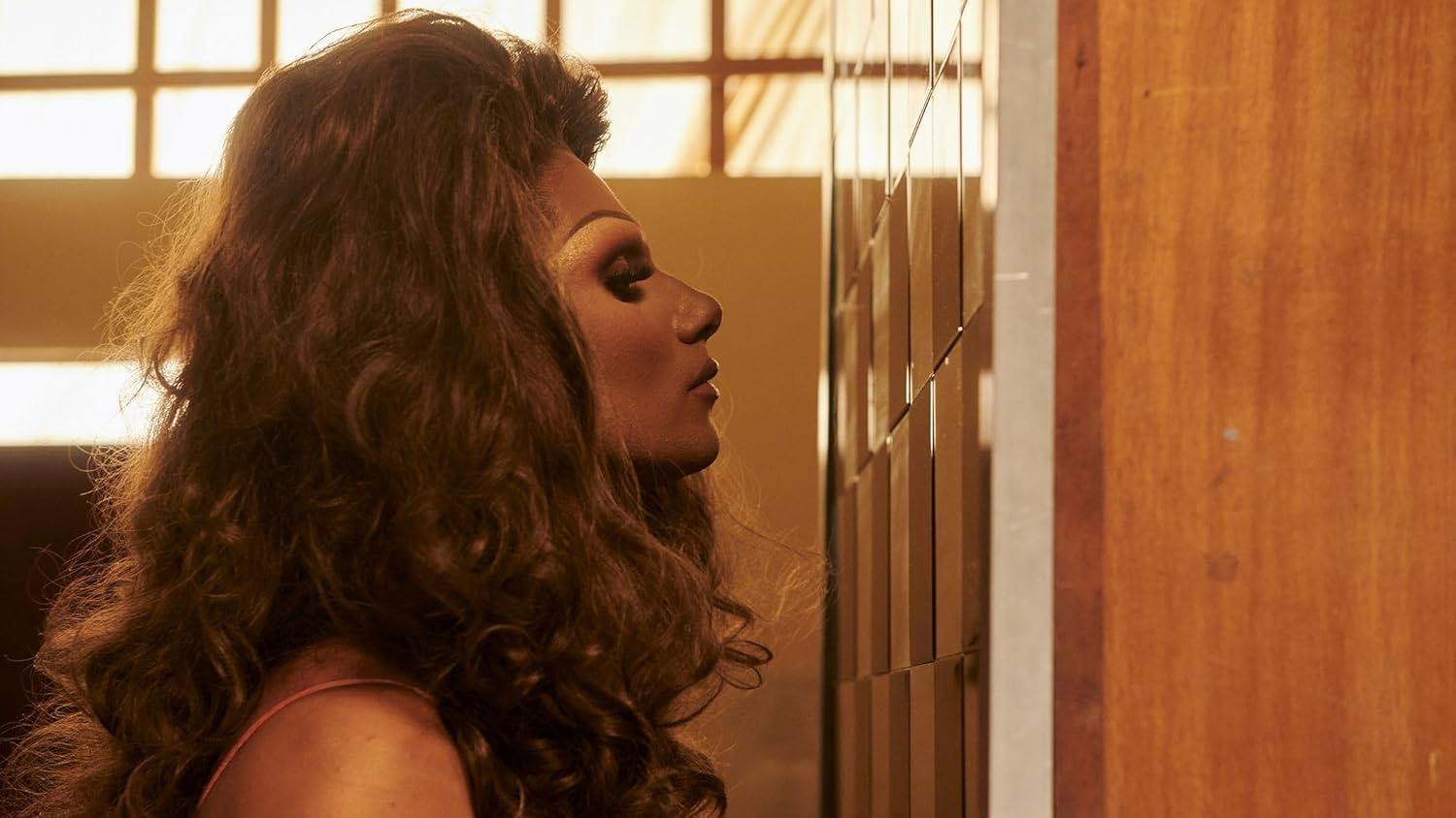 Eine Person mit Make-up und lockigem braunem Haar blickt auf eine geflieste Wand. Der Hintergrund hat ein Fenster mit Gittermuster und Holzwand.