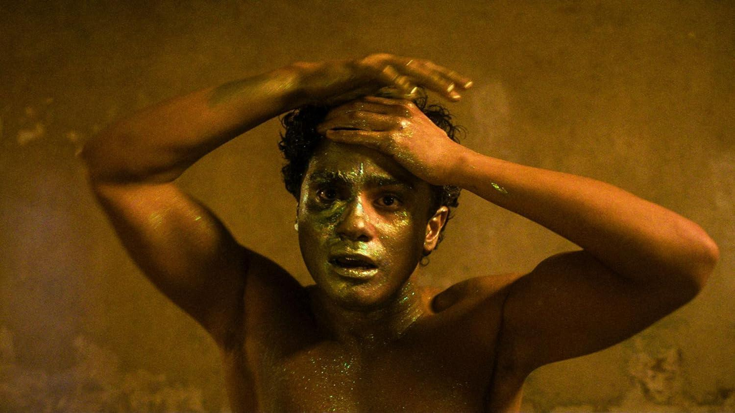 Person mit Goldfarbe im Gesicht und Armen, Hände am Kopf, in schwach beleuchtetem Raum. Hintergrund ist eine warme Farbe.