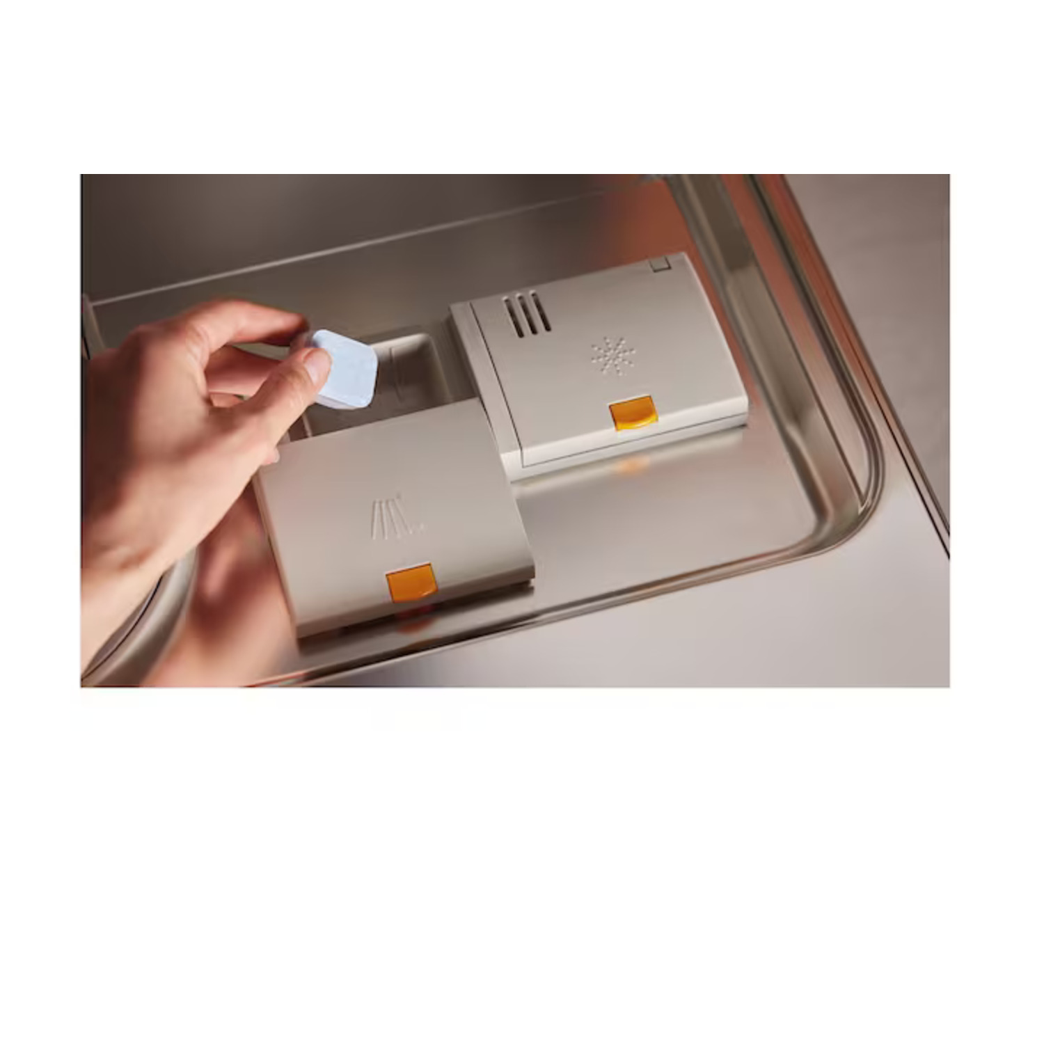 A hand placing a blue tablet into a dishwasher detergent dispenser. Two beige dispenser compartments are visible.