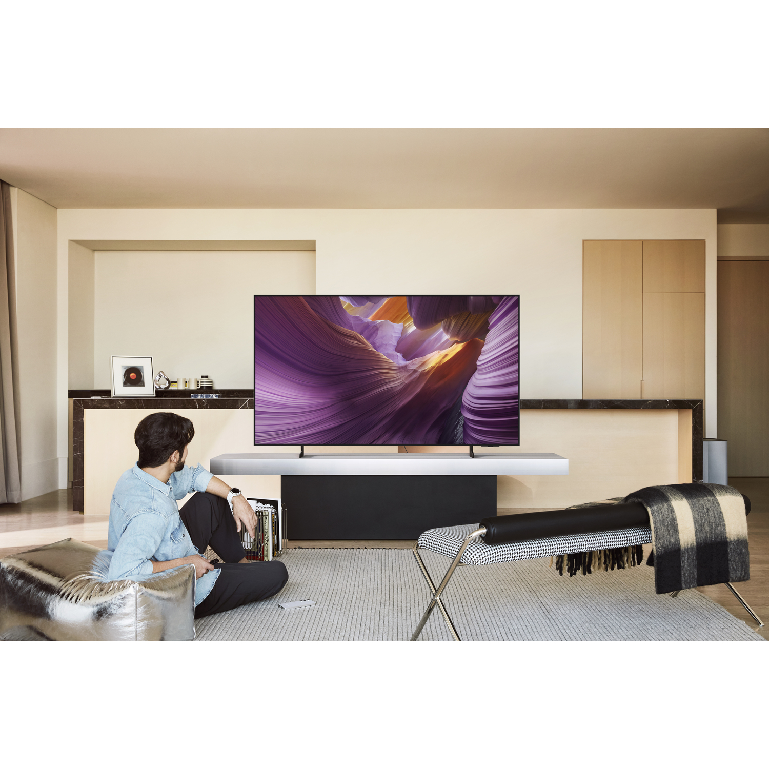 Un homme regarde une grande télévision avec un paysage violet. À droite, un banc avec une couverture. L'homme est sur un pouf argenté.