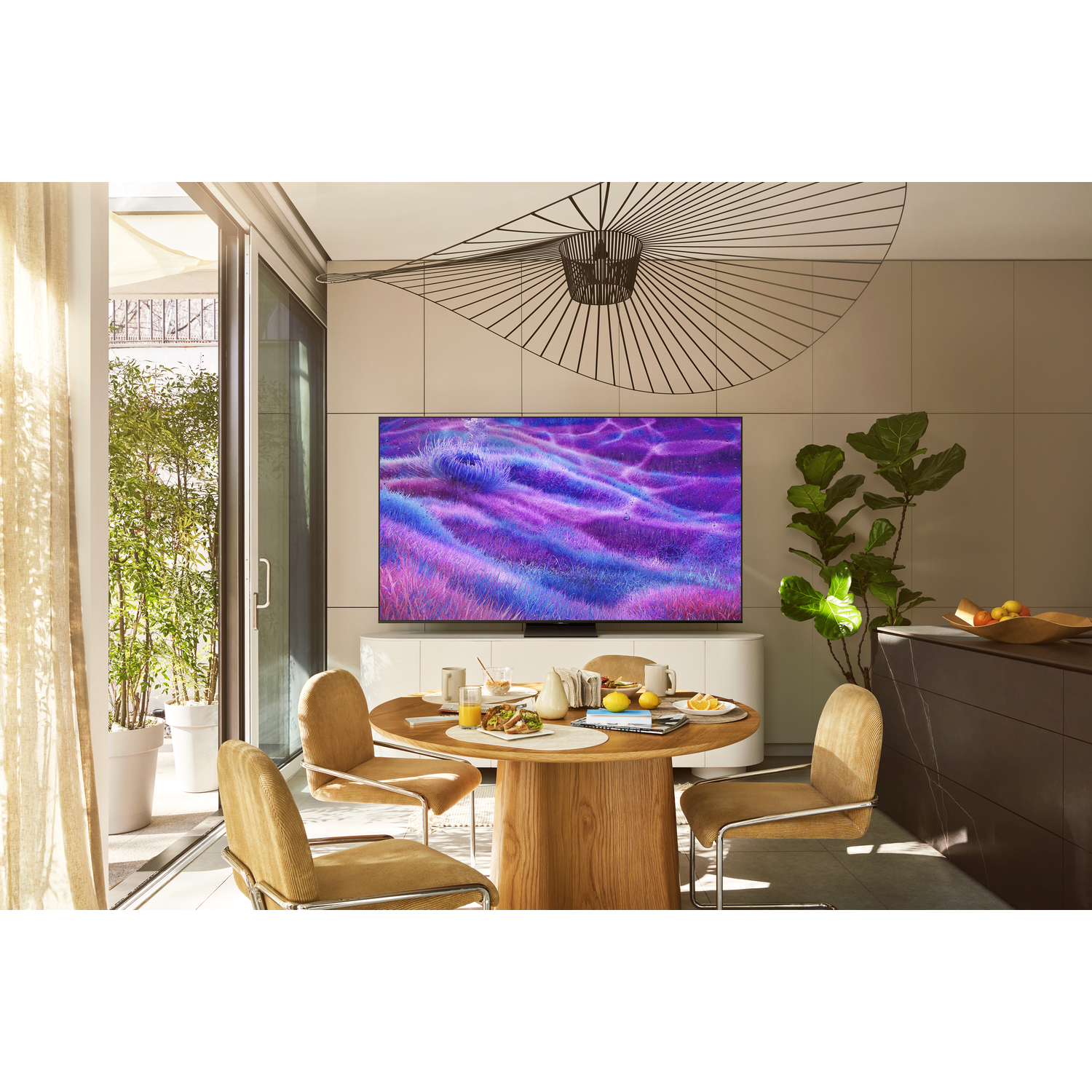 A modern kitchen with a round wooden table set for breakfast. A large TV displays a colorful abstract image. A plant and open door visible.