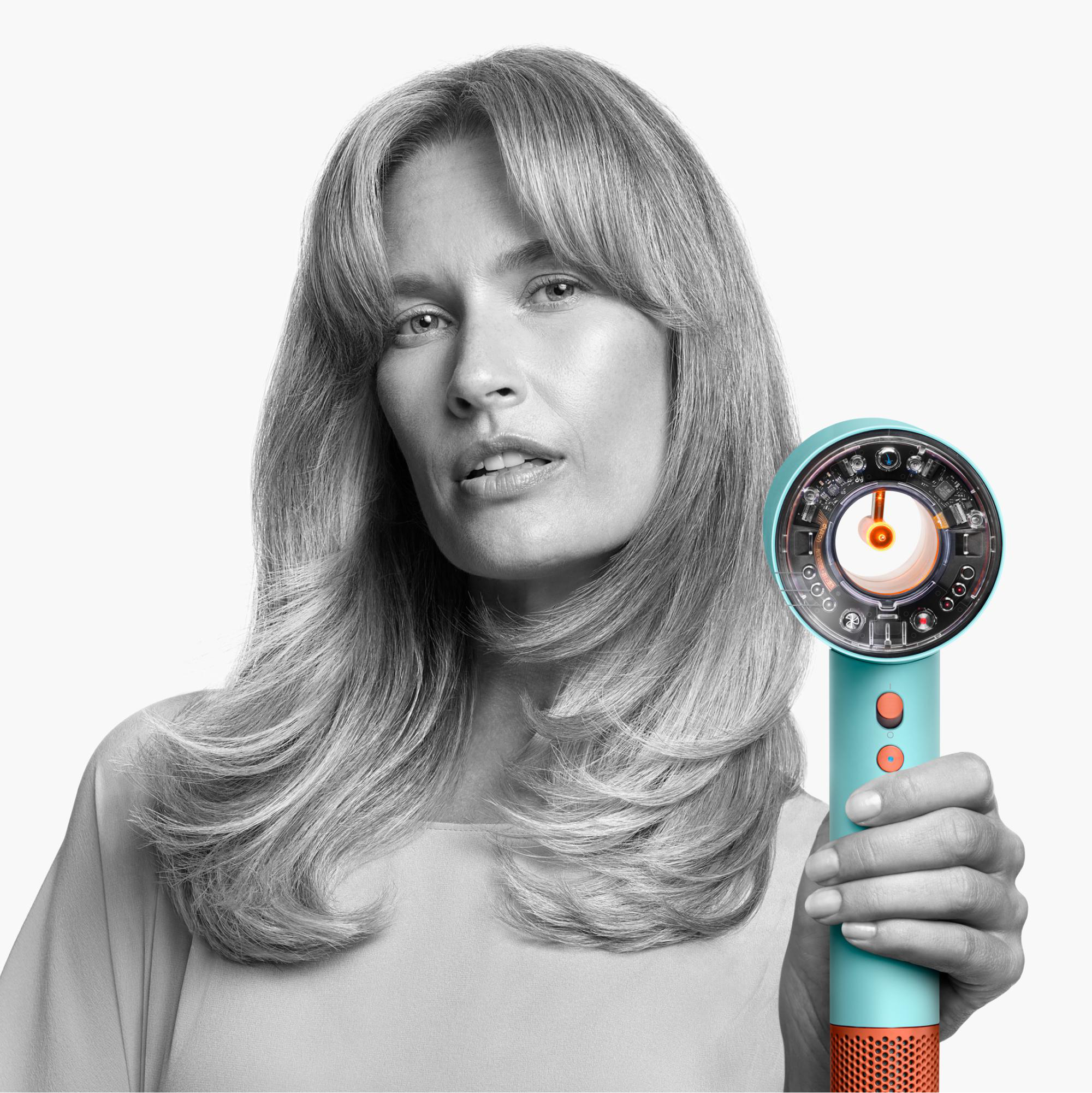 Frau hält einen türkis-orangenen Föhn, blickt in die Kamera. Ihr blondes Haar ist lang und wellig, vor weißem Hintergrund.