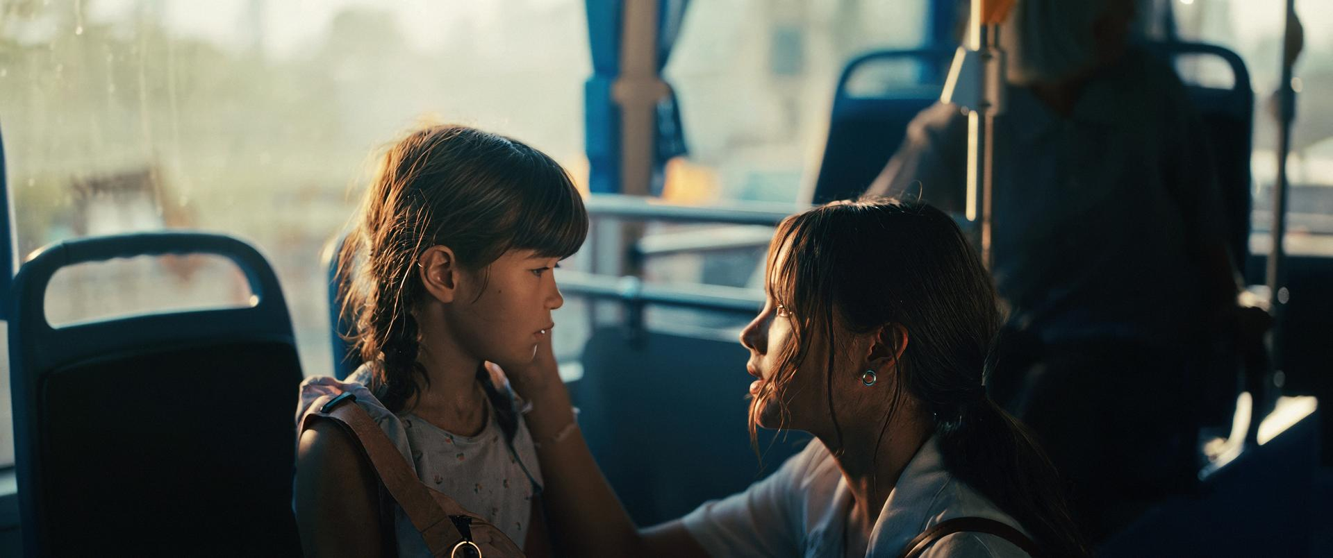 Ein junges Mädchen und eine Frau sitzen sich in einem Bus gegenüber. Die Frau berührt das Gesicht des Mädchens. Sonnenlicht scheint durch das Fenster.