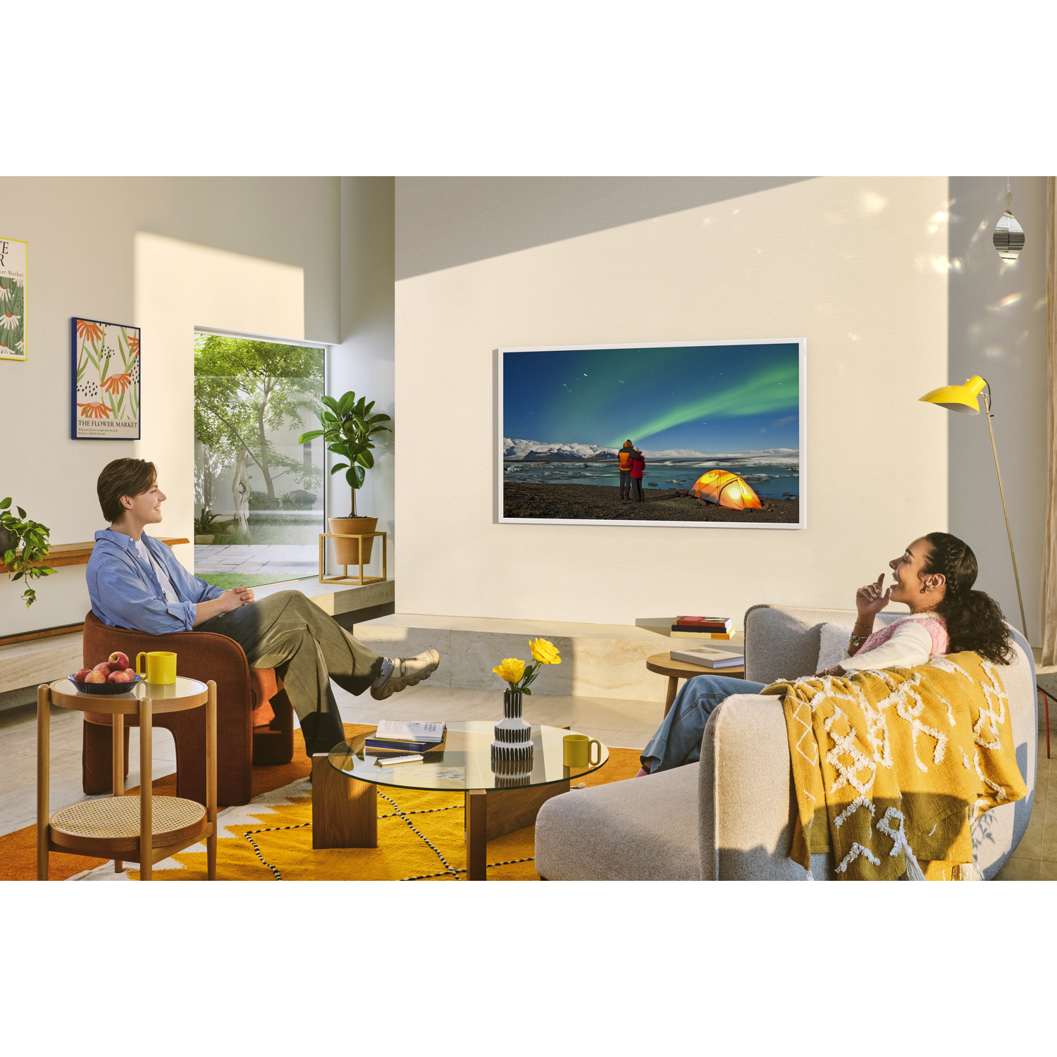 Two people relax in a living room, one on a chair, the other on a sofa. A TV displays a scenic view. Yellow accents and a window are visible.