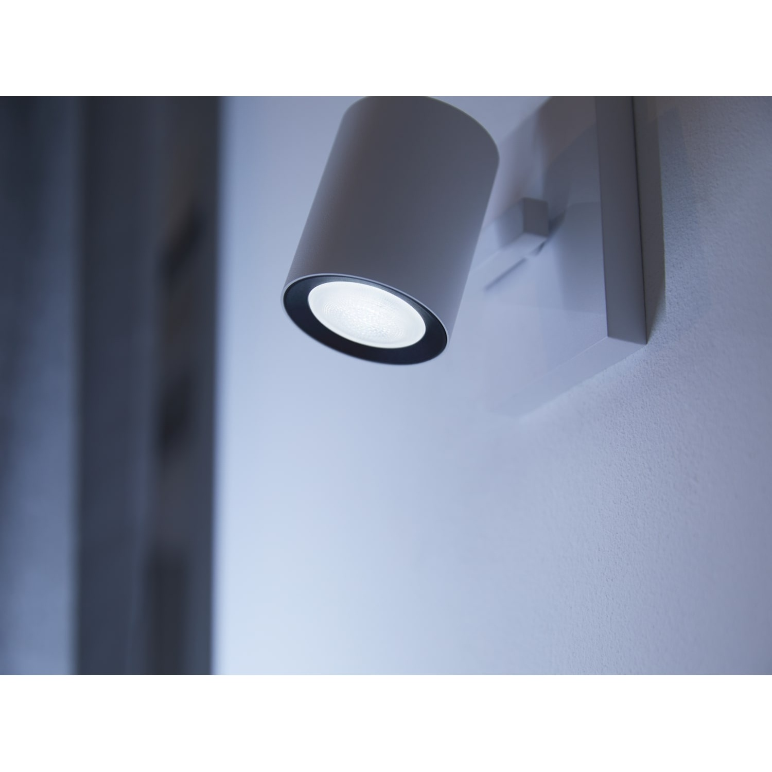 Un faretto cilindrico bianco è montato su un muro bianco. La luce è accesa, illuminando la parete. Uno sfondo sfocato suggerisce una stanza.